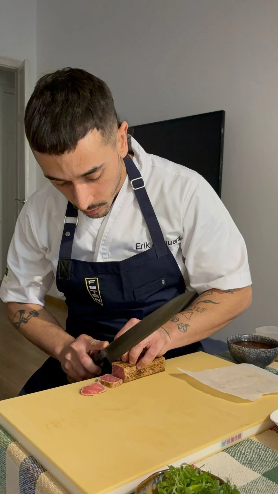 Un hombre con delantal negro y camiseta blanca cortando carne con un cuchillo en una tabla de cortar amarilla en una cocina.