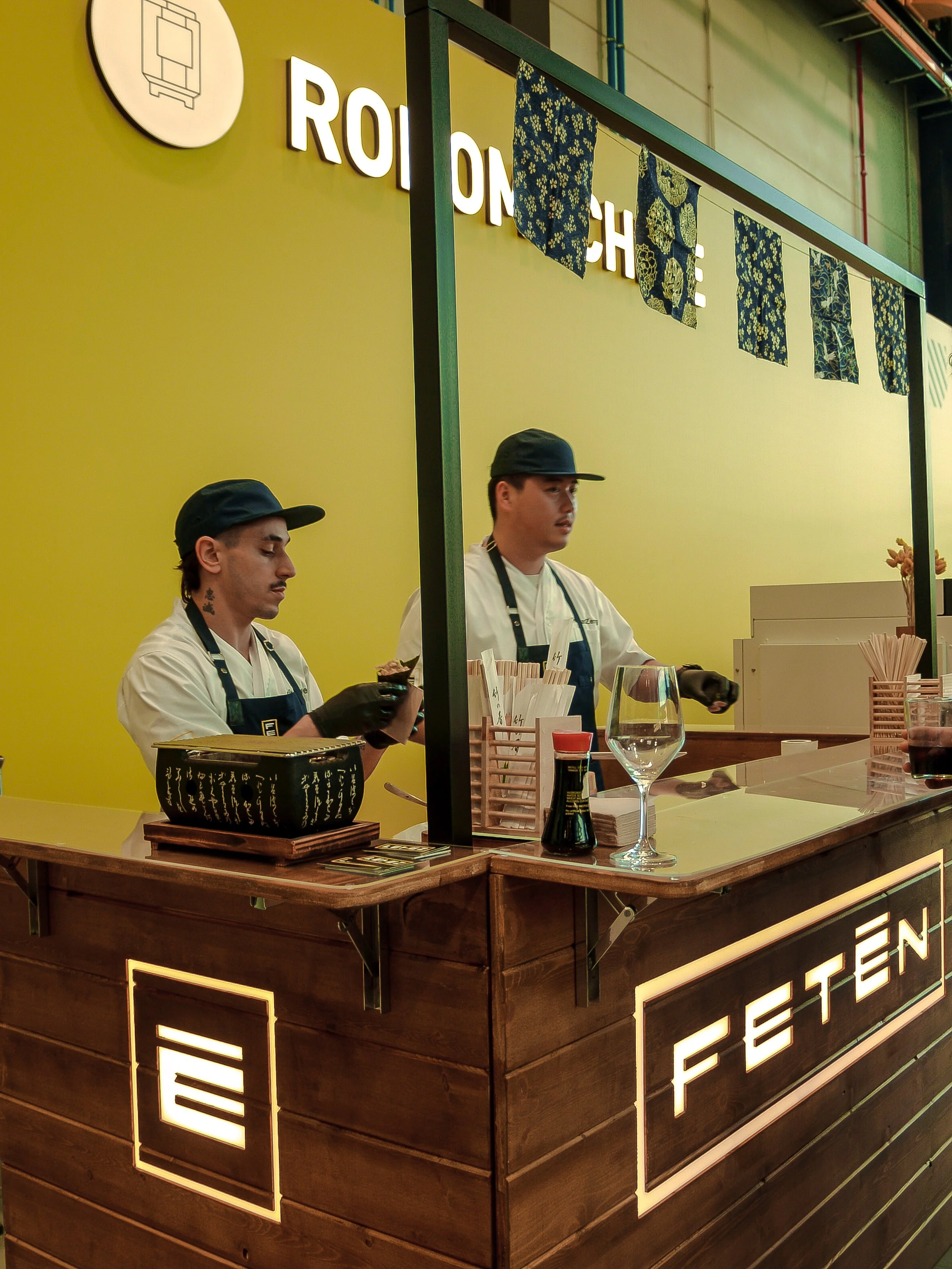 Dos cocineros en una cocina de estilo japonés en una feria o evento, con decoración en tonos oscuros y luces cálidas.