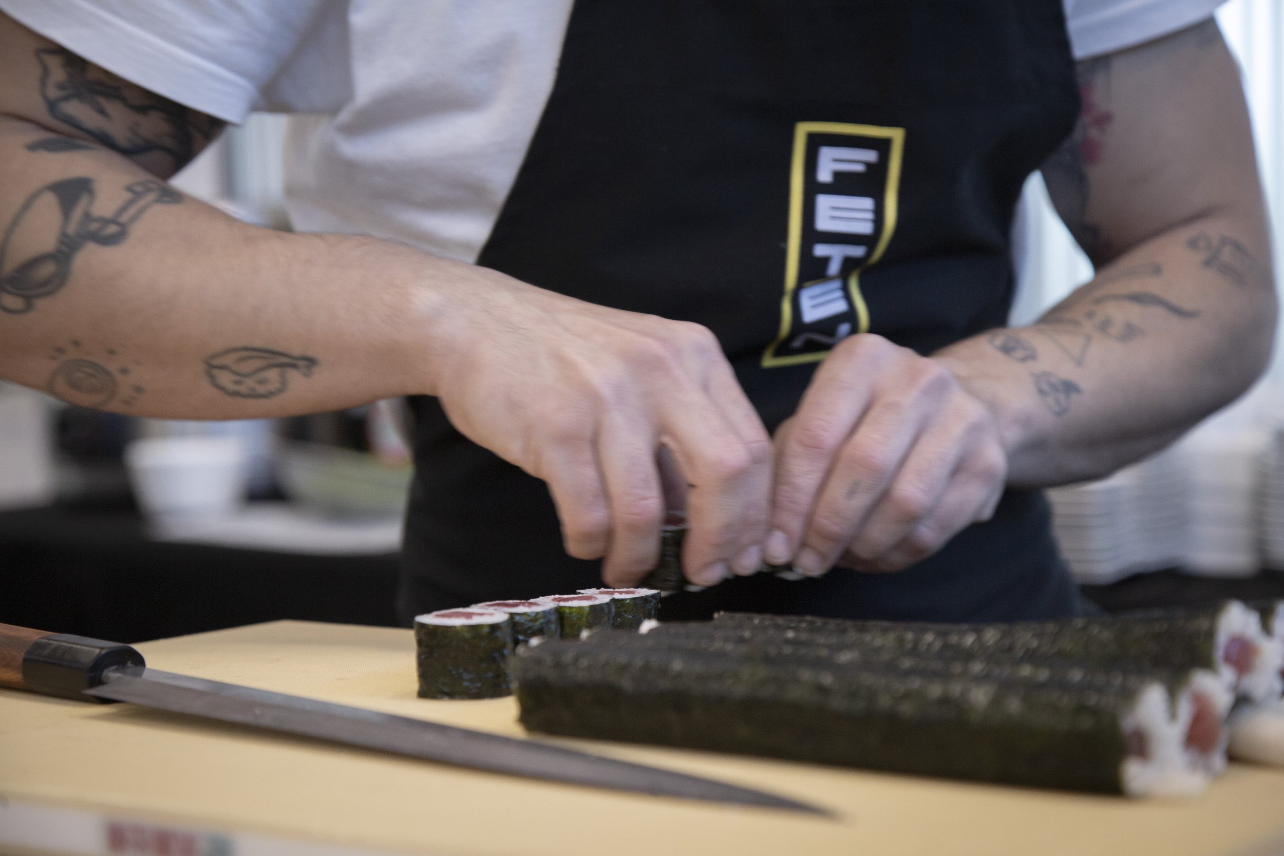 Persona cortando sushi sobre una mesa de madera, con tatuajes visibles en los brazos.