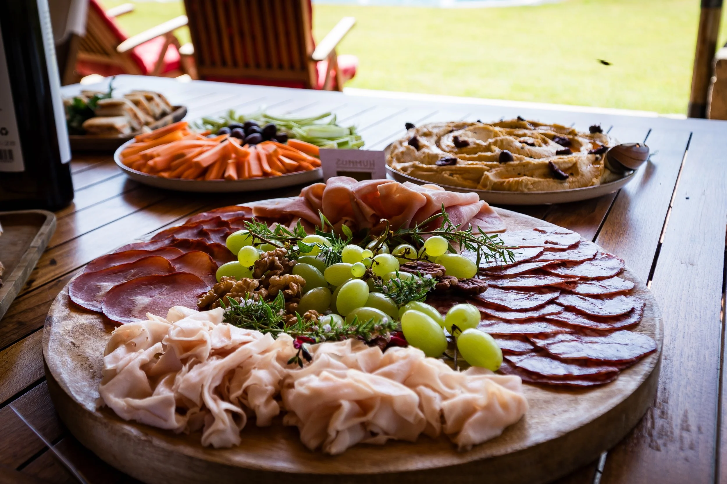 Tabla con embutidos, queso, uvas, nueces, verduras, y una pizza en un ambiente al aire libre.