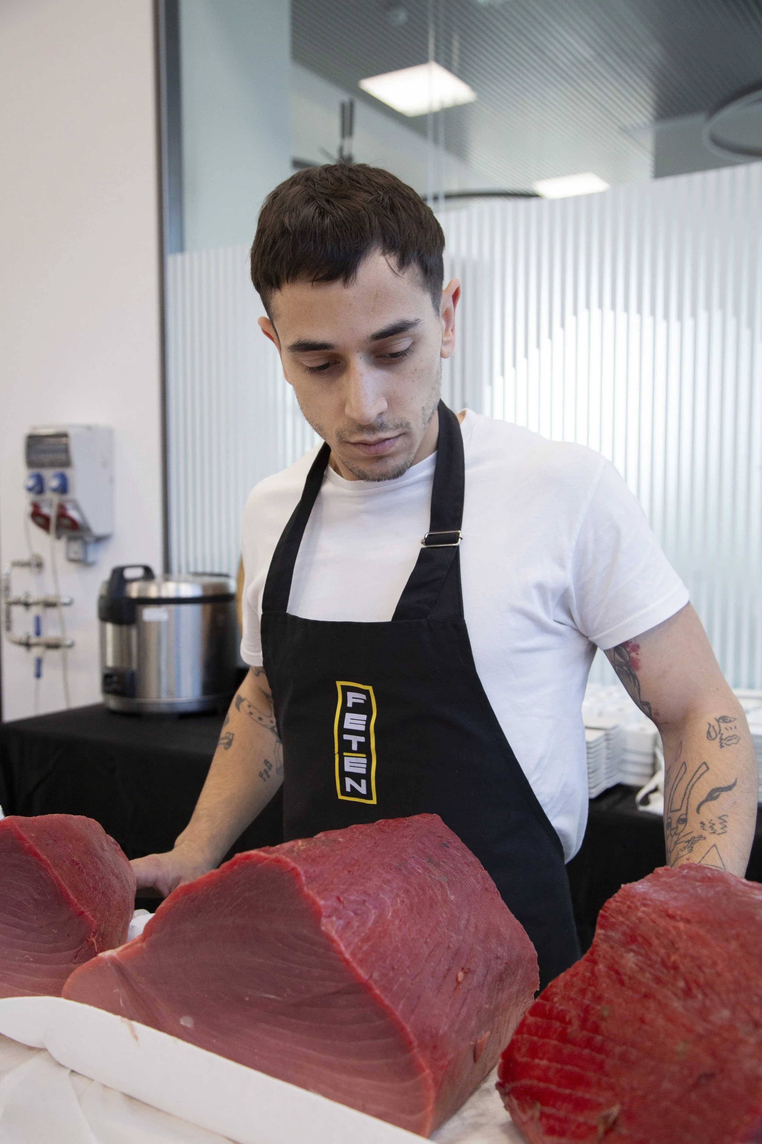 Joven hombre con tatuajes en los brazos, usando camiseta blanca y delantal negro, preparando carne en una cocina moderna.