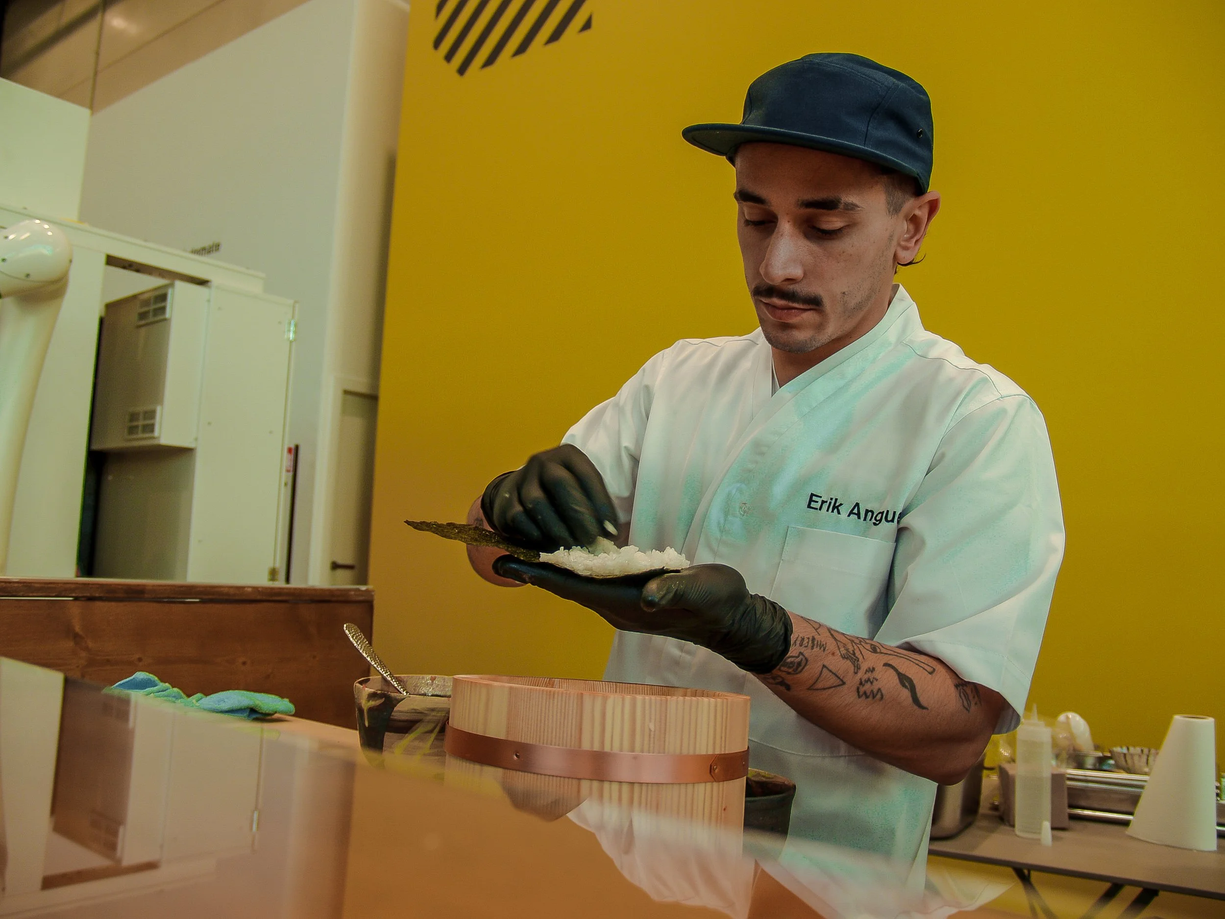 Joven cocinero con gorra negra y camiseta blanca, con tatuajes en brazos, sirviendo comida en un recipiente de madera en una cocina moderna con fondo amarillo.