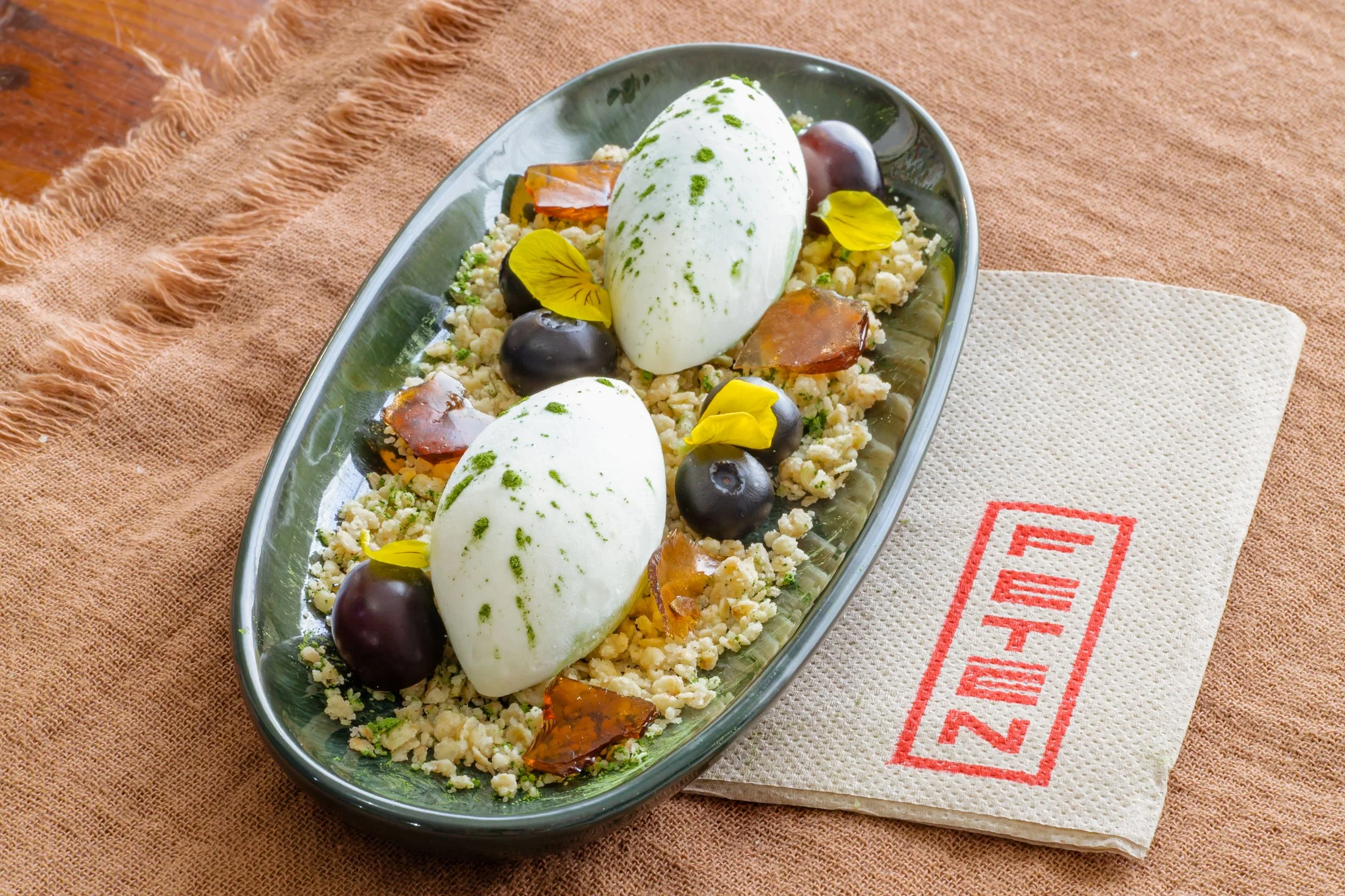 Plato de ensalada con queso fresco, aceitunas negras, hojas de albaca y flores comestibles, sobre una mesa de tela color tierra, con un servilleta de papel con letras rojas.