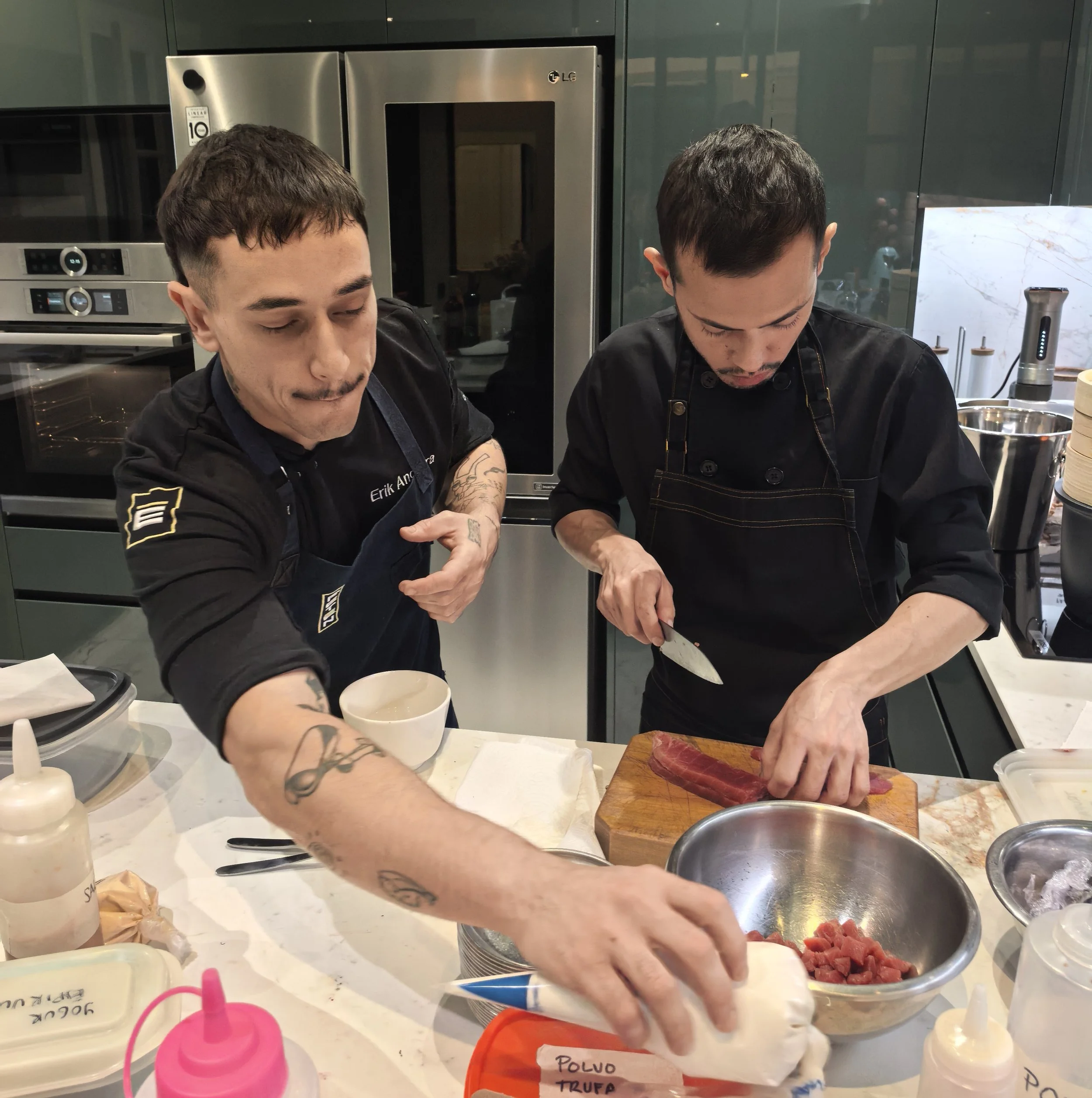 Dos hombres en una cocina, uno de ellos cortando carne roja en una tabla, mientras el otro observa. La encimera tiene varios ingredientes y utensilios, y en el fondo hay electrodomésticos de acero inoxidable y gabinetes verdes.