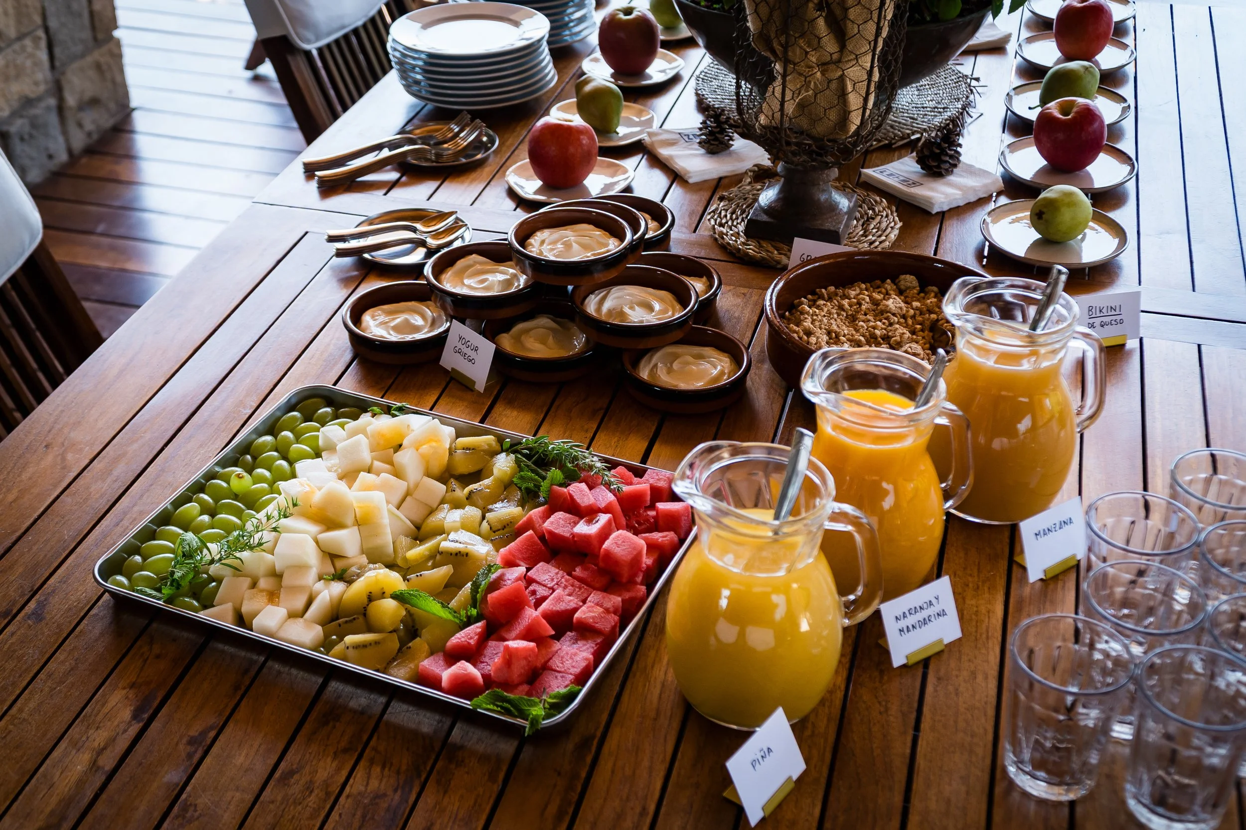 Mesa de madera con variedad de frutas y desayunos, incluyendo yogur griego, frutas cortadas, jugos y platos con manzana, piña y sandía, en un entorno de comida en interior con luz natural.