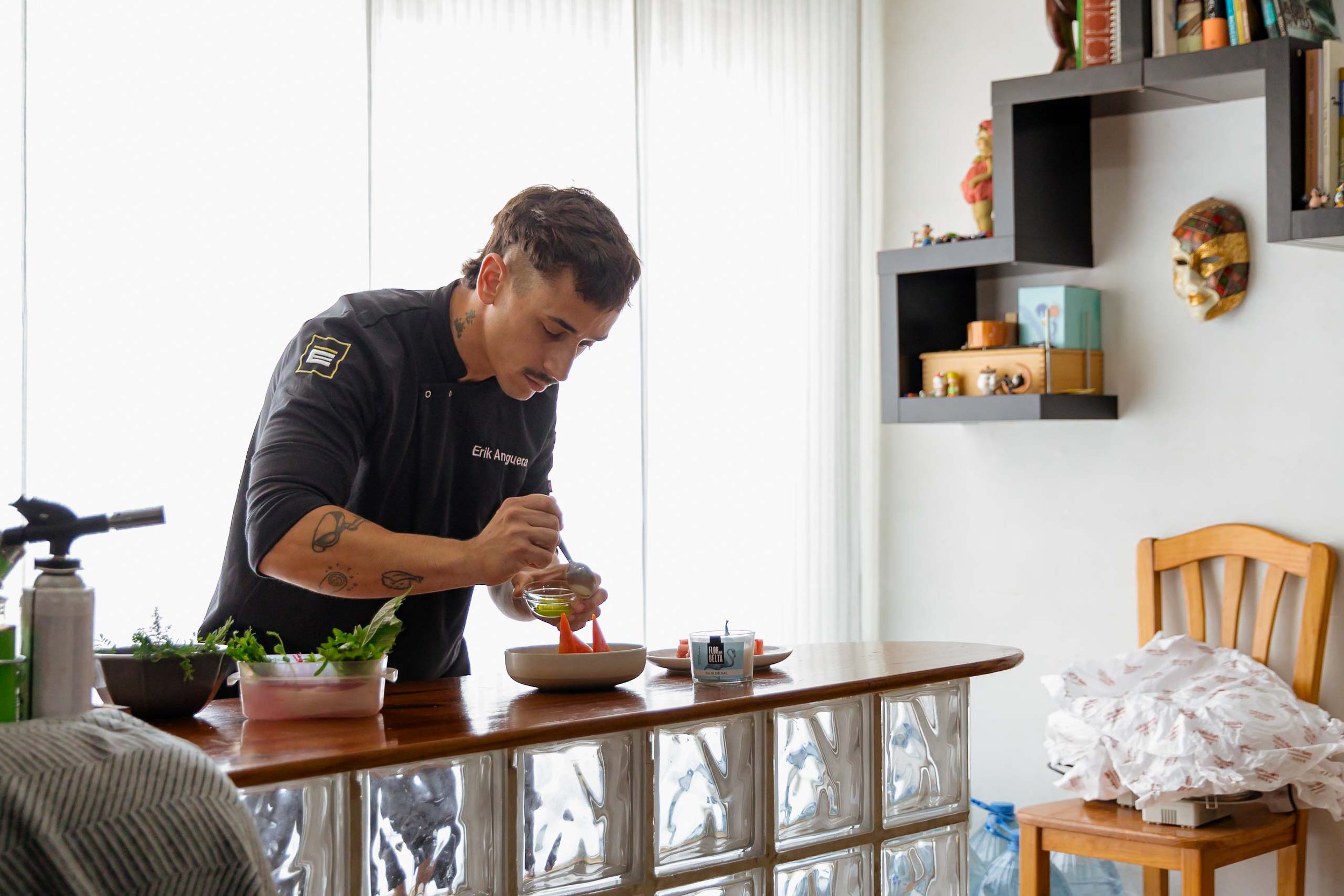 Hombre preparando comida en una mesa en una cocina luminosa, decorada con estanterías y máscaras artísticas.