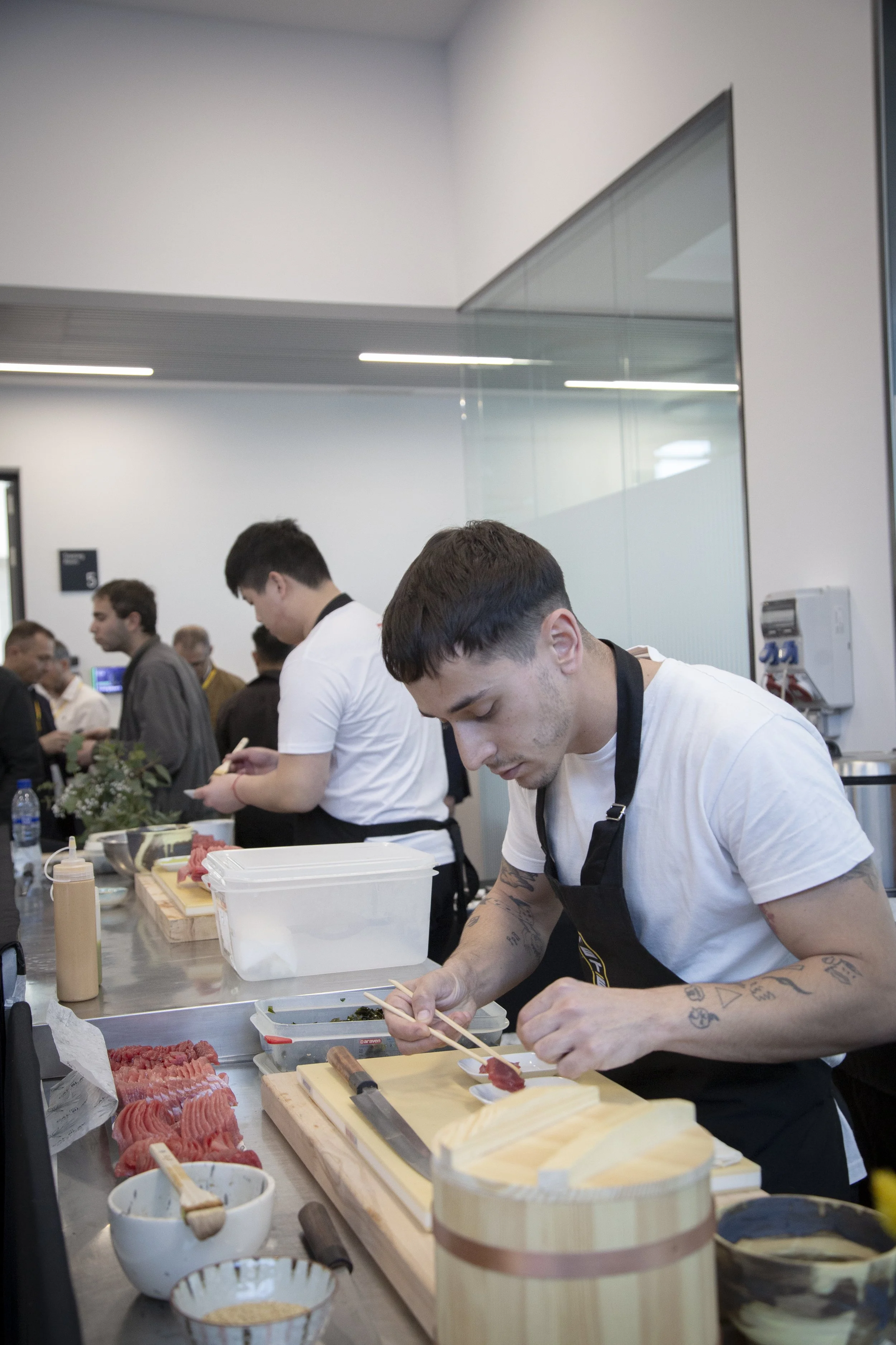 Joven cocinero preparando sushi en una cocina industrial, rodeado de ingredientes y utensilios de cocina.