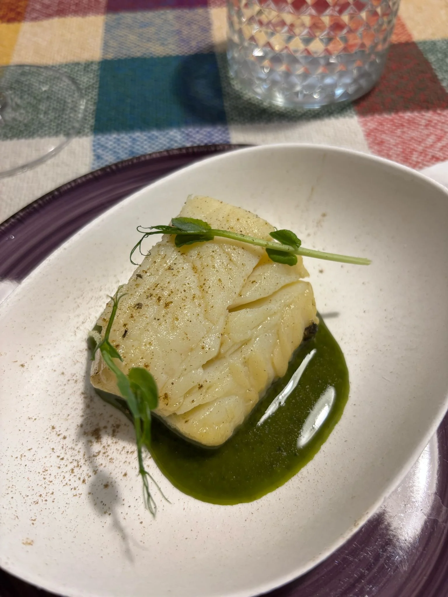 Filete de bacalao confitado sobre plato blanco con salsa verde, decorado con brotes verdes y polvo de especias, sobre mantel de cuadros de colores.