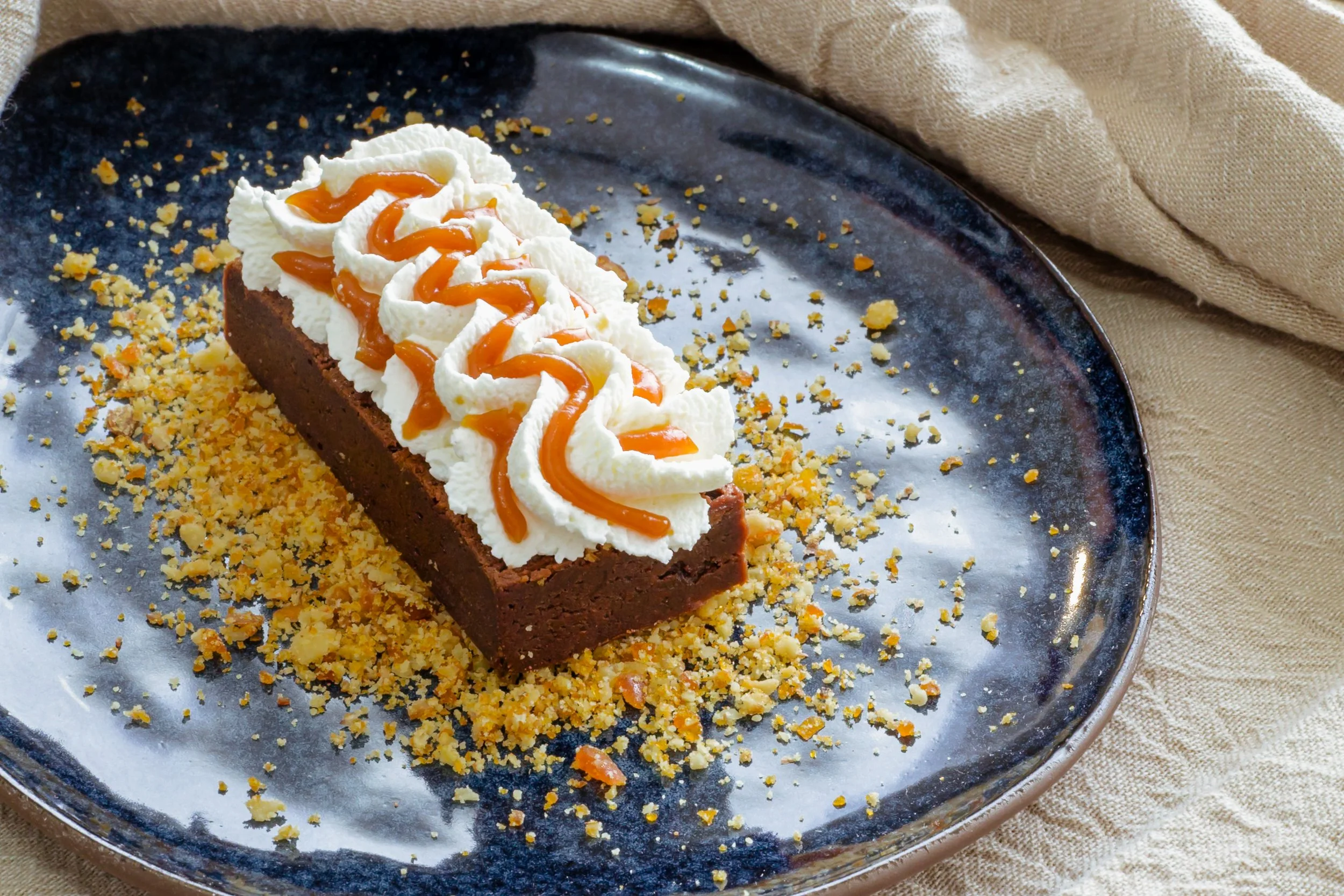 Porción de tarta de chocolate decorada con crema y salsa de caramelo, presentada en plato de metal, con migajas de galleta alrededor y paño de tela beige de fondo.