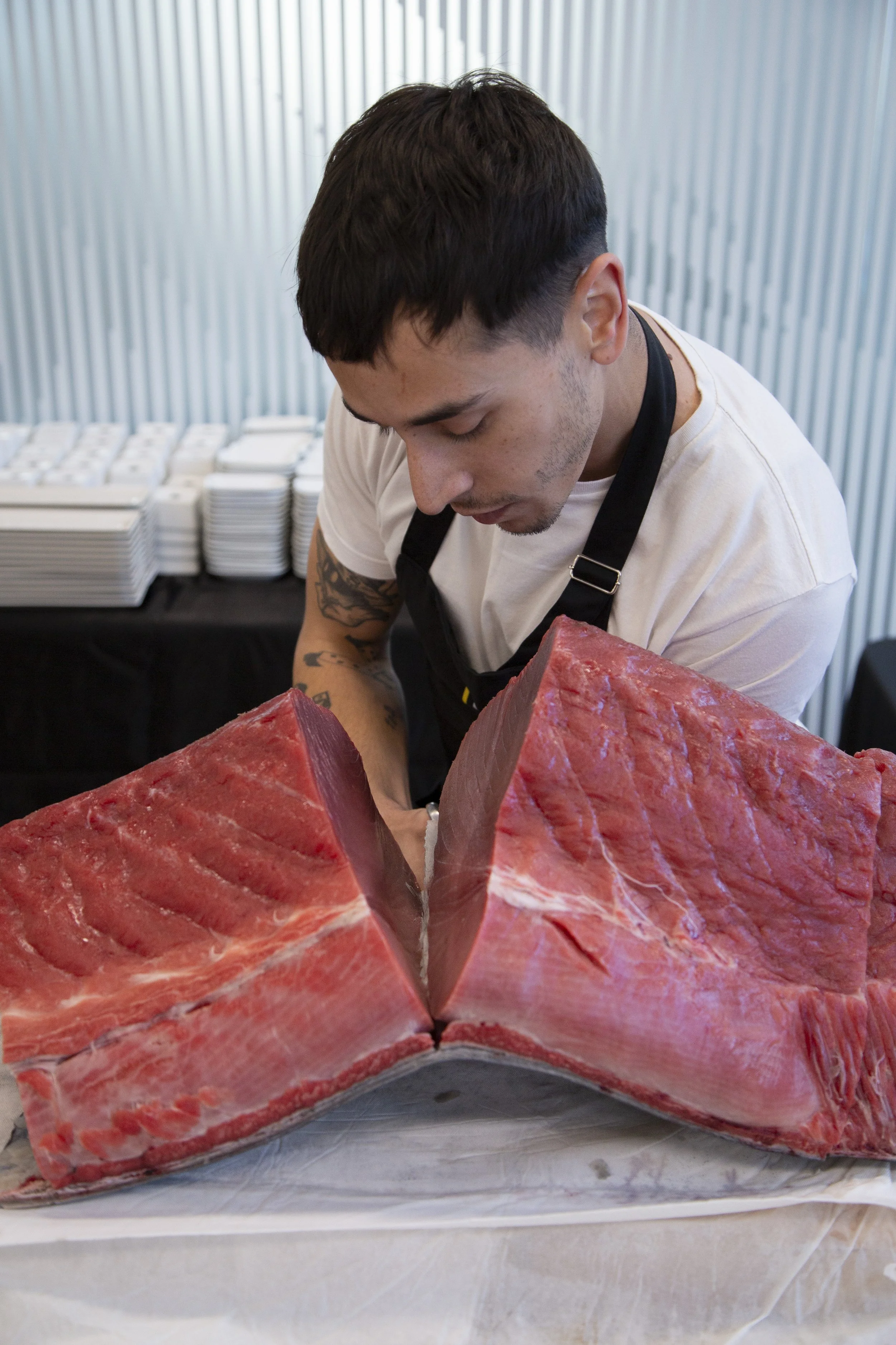 Un hombre con cabello oscuro, camiseta blanca y delgada, y tatuajes en su brazo, trabaja con un bloque de carne en un ambiente de cocina o carnicería.