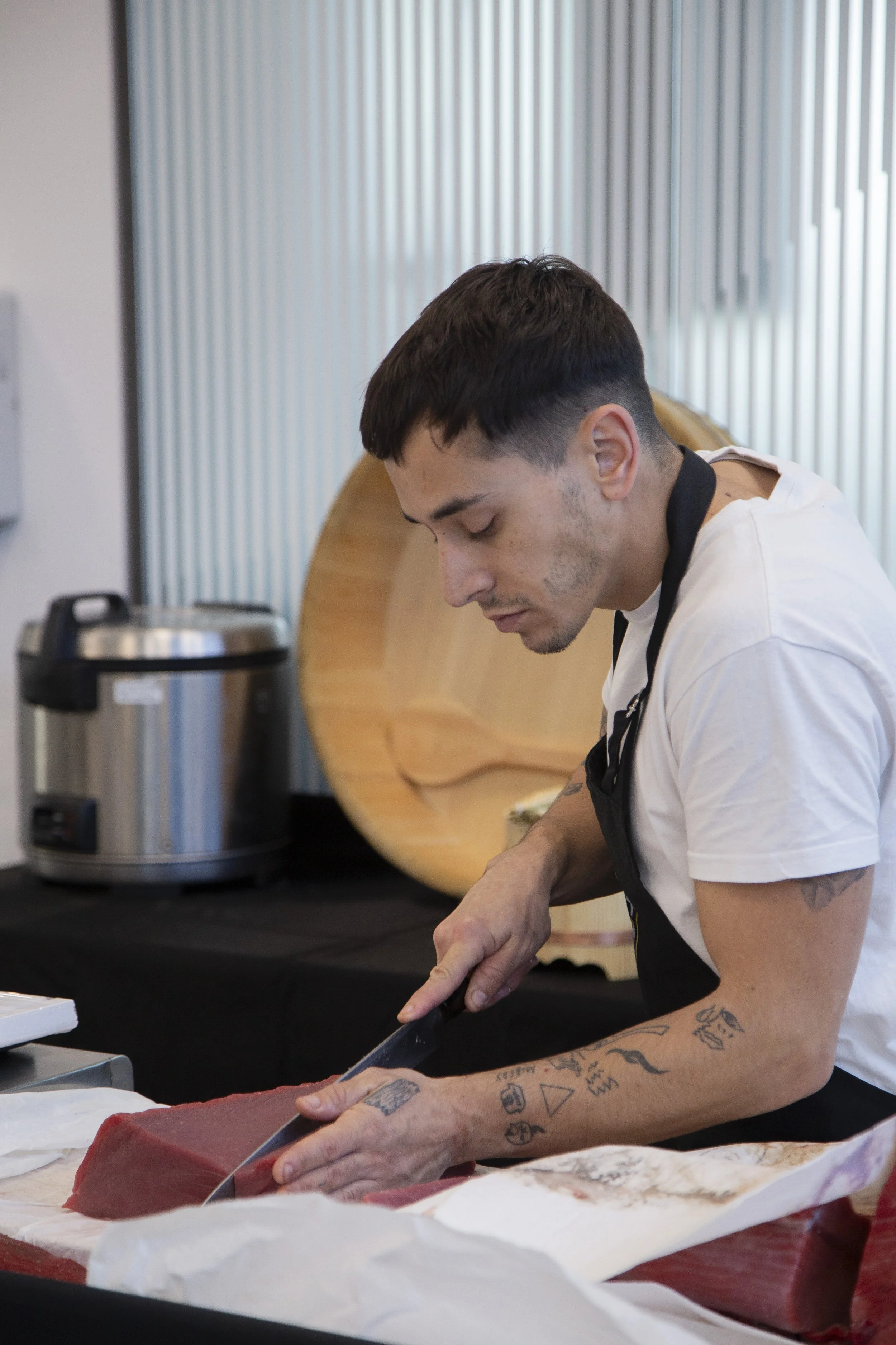 Un joven chef con tatuajes en los brazos, cortando carne en una cocina moderna.