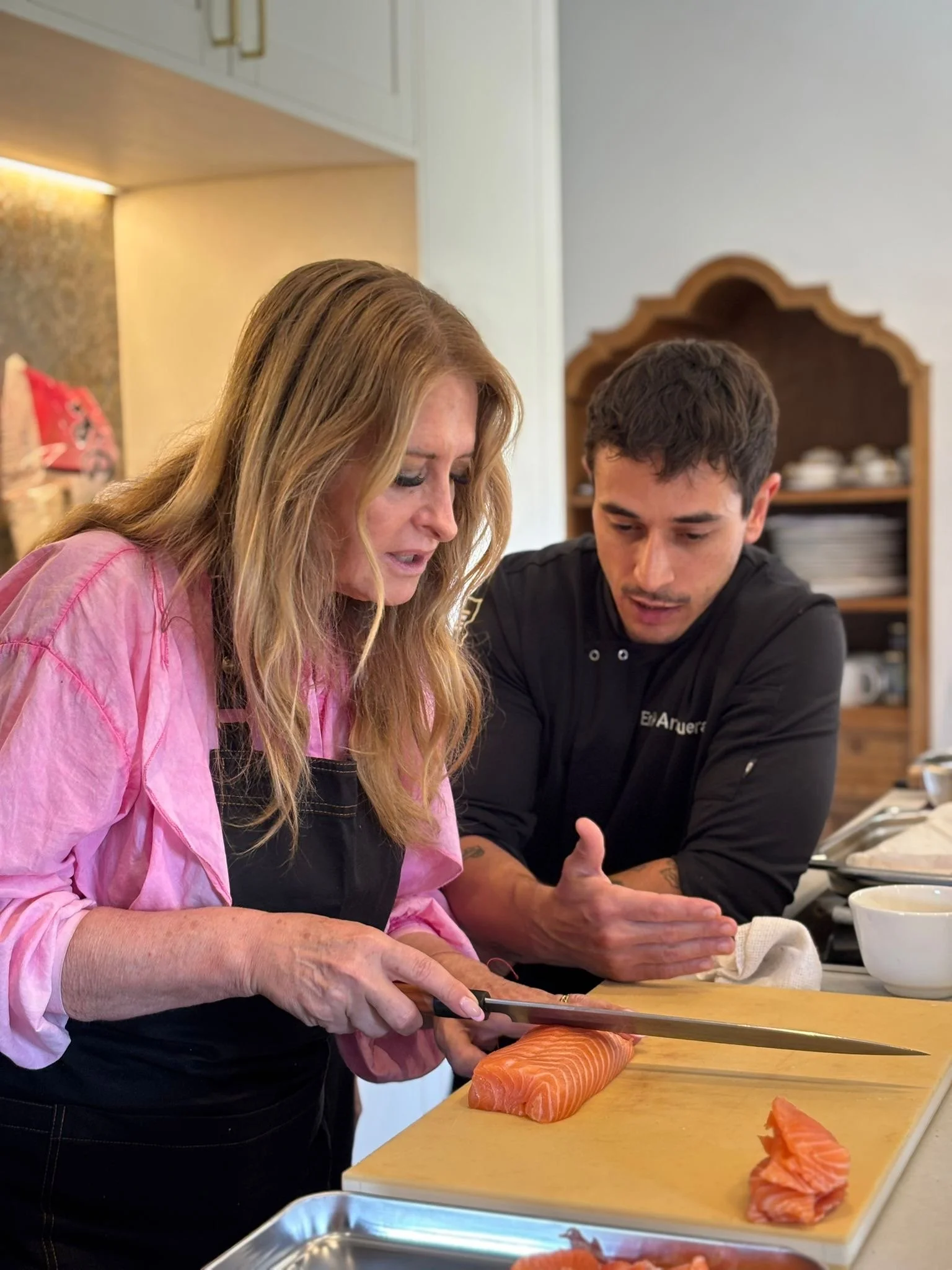 Una mujer y un hombre en la cocina están cortando salmón en una tabla de cortar. La mujer viste una blusa rosa y el hombre viste una camiseta negra. Al fondo hay estanterías con platos y otros utensilios de cocina.