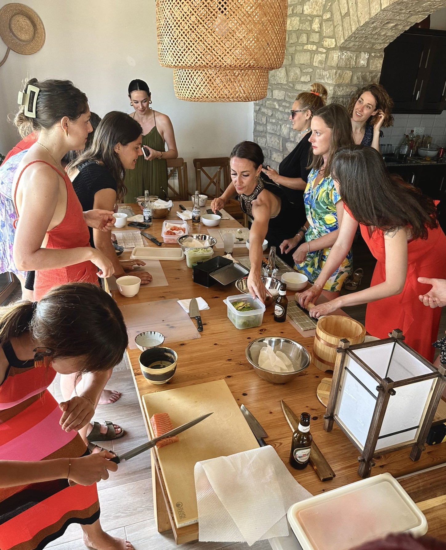 Varias mujeres en una cocina o comedor, participando en un taller de sushi, en torno a una mesa con ingredientes y utensilios de cocina.