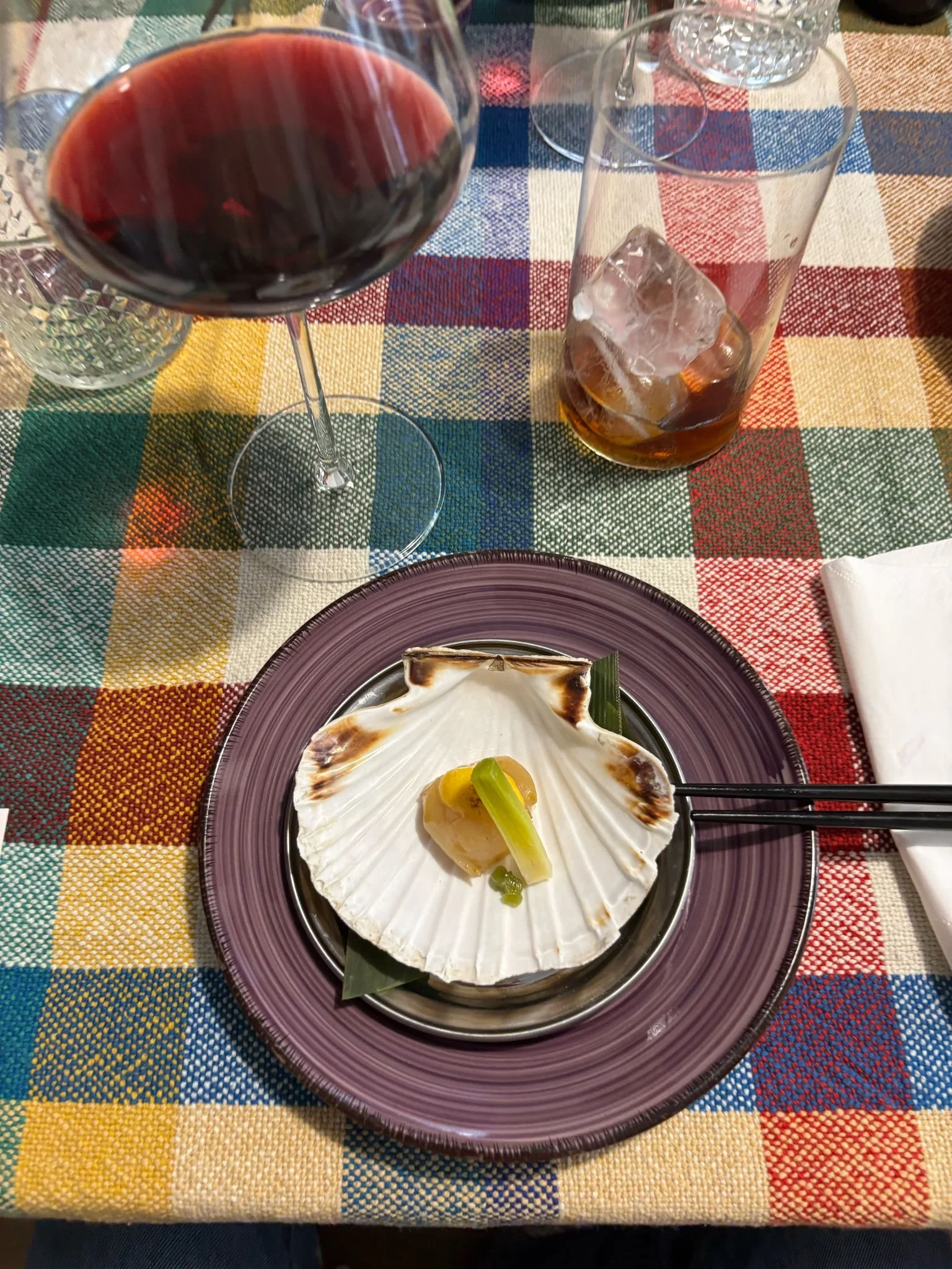 Plato de mariscos con concha, limón y cebolla, acompañado de un vaso de bebida con hielo y un vaso de vino tinto en una mesa con mantel colorido.