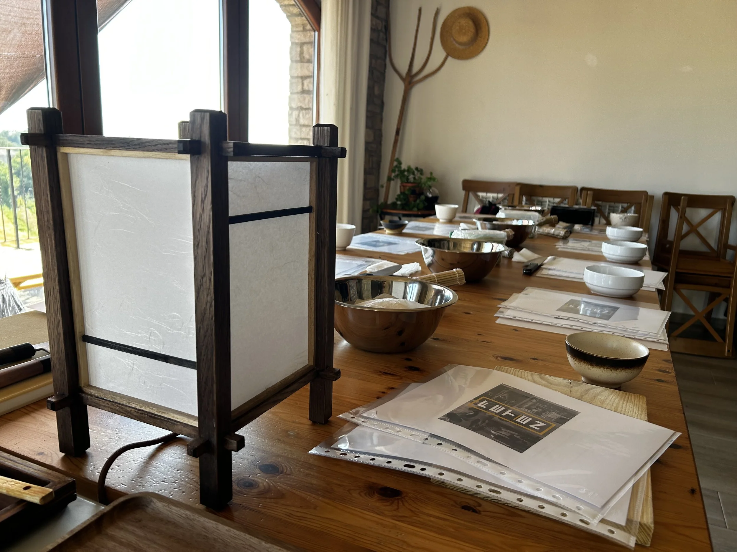 Mesa de madera con varios instrumentos y utensilios de cocina, y un separador de papel en primer plano, en una habitación con ventana y paredes decoradas con paja y plantas.