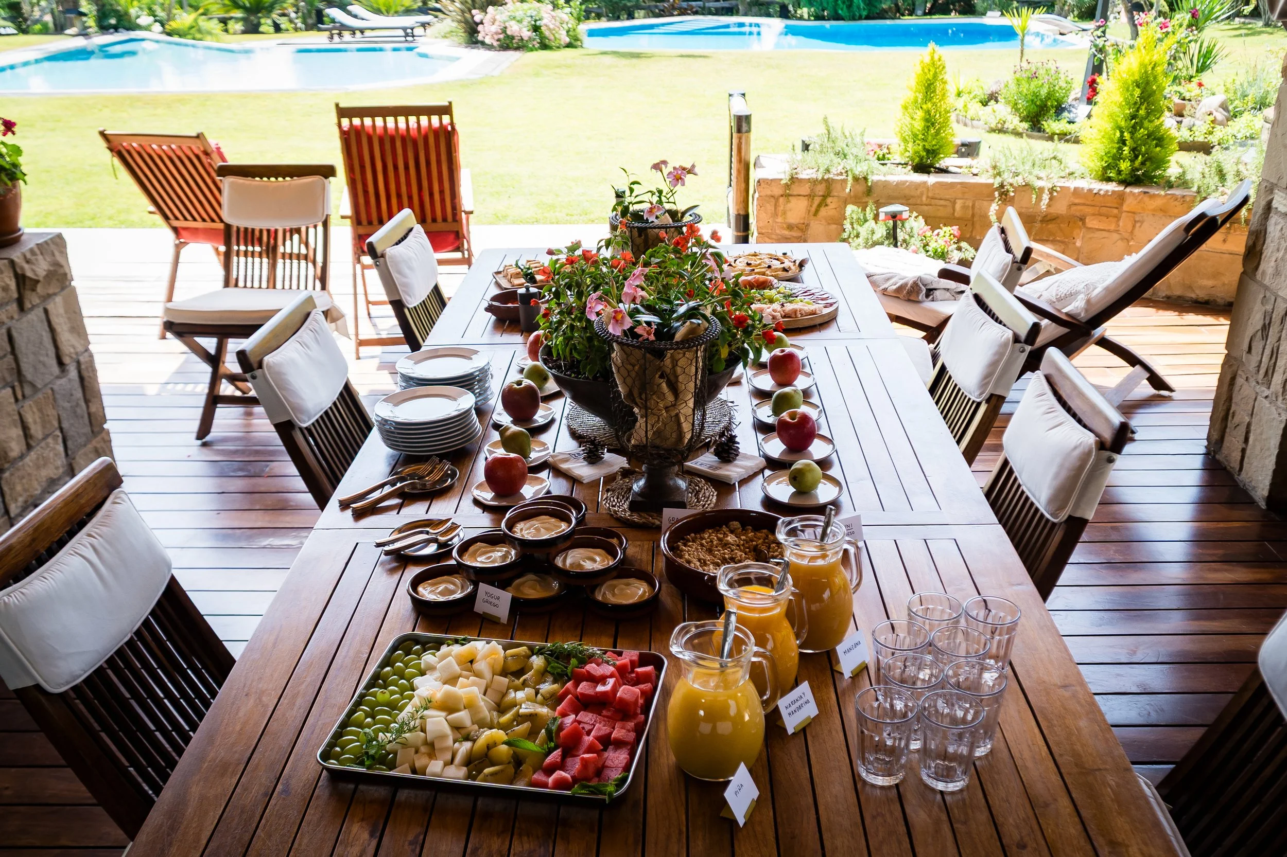 Desayuno al aire libre en una terraza con vista a un jardín con albercas. Mesa con frutas, yogurt, jugos y otros alimentos, rodeada de sillas con cojines.