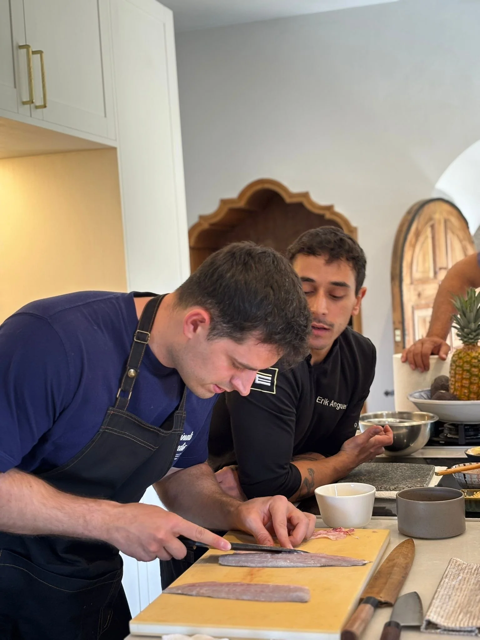 Dos hombres cocinando en una cocina moderna. Uno de ellos está picando un pescado en una tabla de madera, ambos llevan delantales y hay varios utensilios y ingredientes sobre la encimera.
