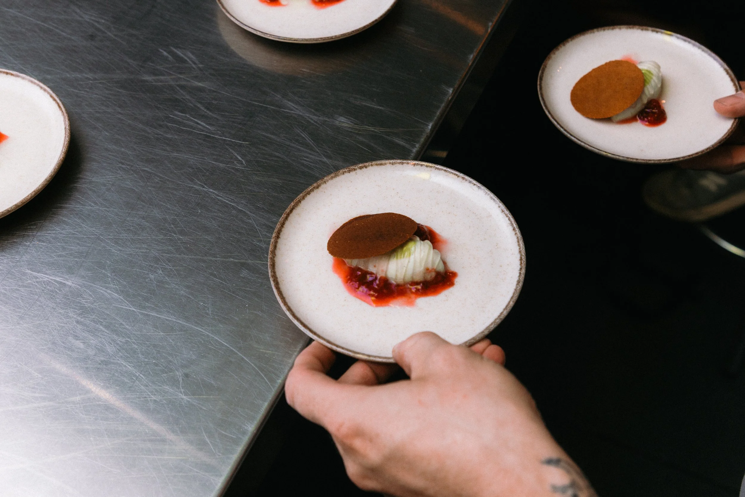 Mano sosteniendo un pequeño plato con postre que tiene un obleas de color marrón, crema blanca, fruta roja y salsa.