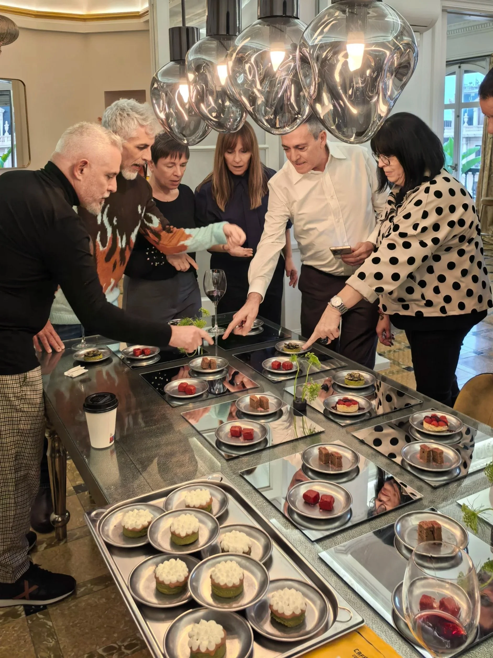 Varias personas observan y discuten una selección de postres en una mesa de presentación, con platos que contienen diferentes tipos de pasteles y postres. Hay un decorado elegante en el ambiente y grandes lámparas modernas en el techo.