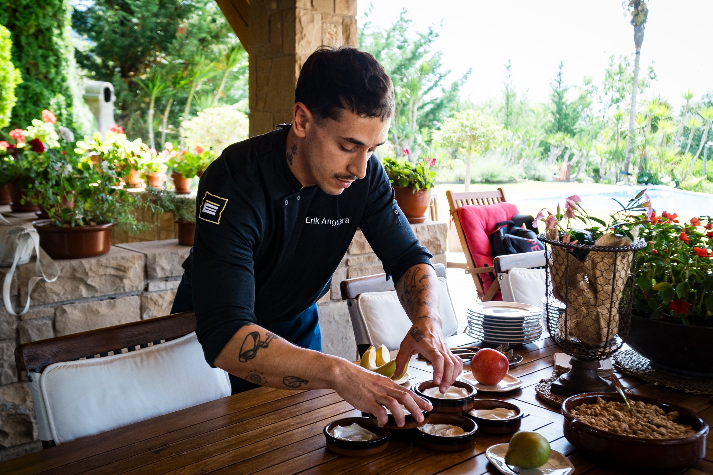 Hombre sirviendo comida en una mesa de madera en un espacio abierto con paredes de piedra y muchas plantas verdes. Chef privado para teambuildings.