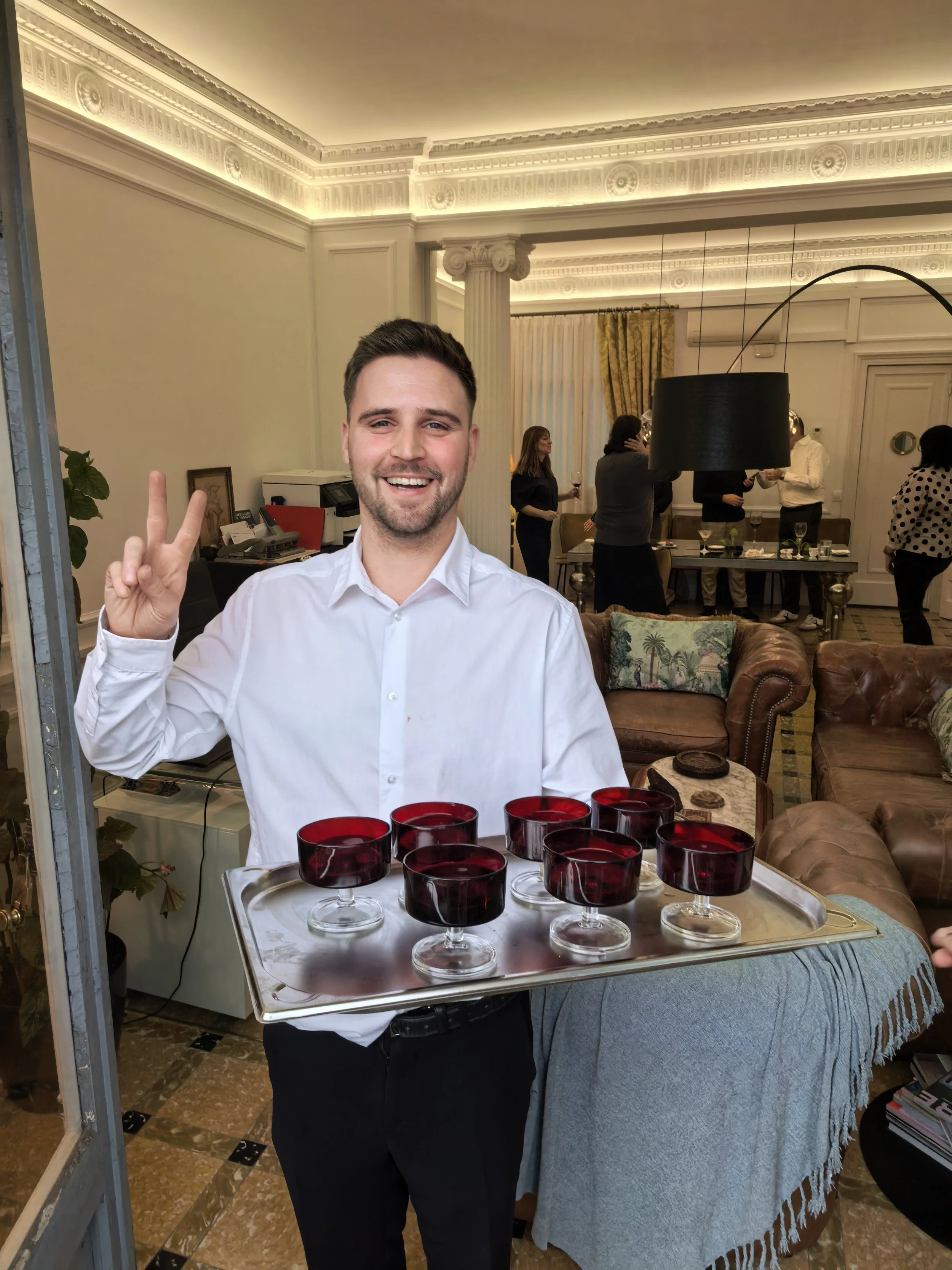 Hombre sonriente con camisa blanca sosteniendo una bandeja con varias copas de vino tinto.
