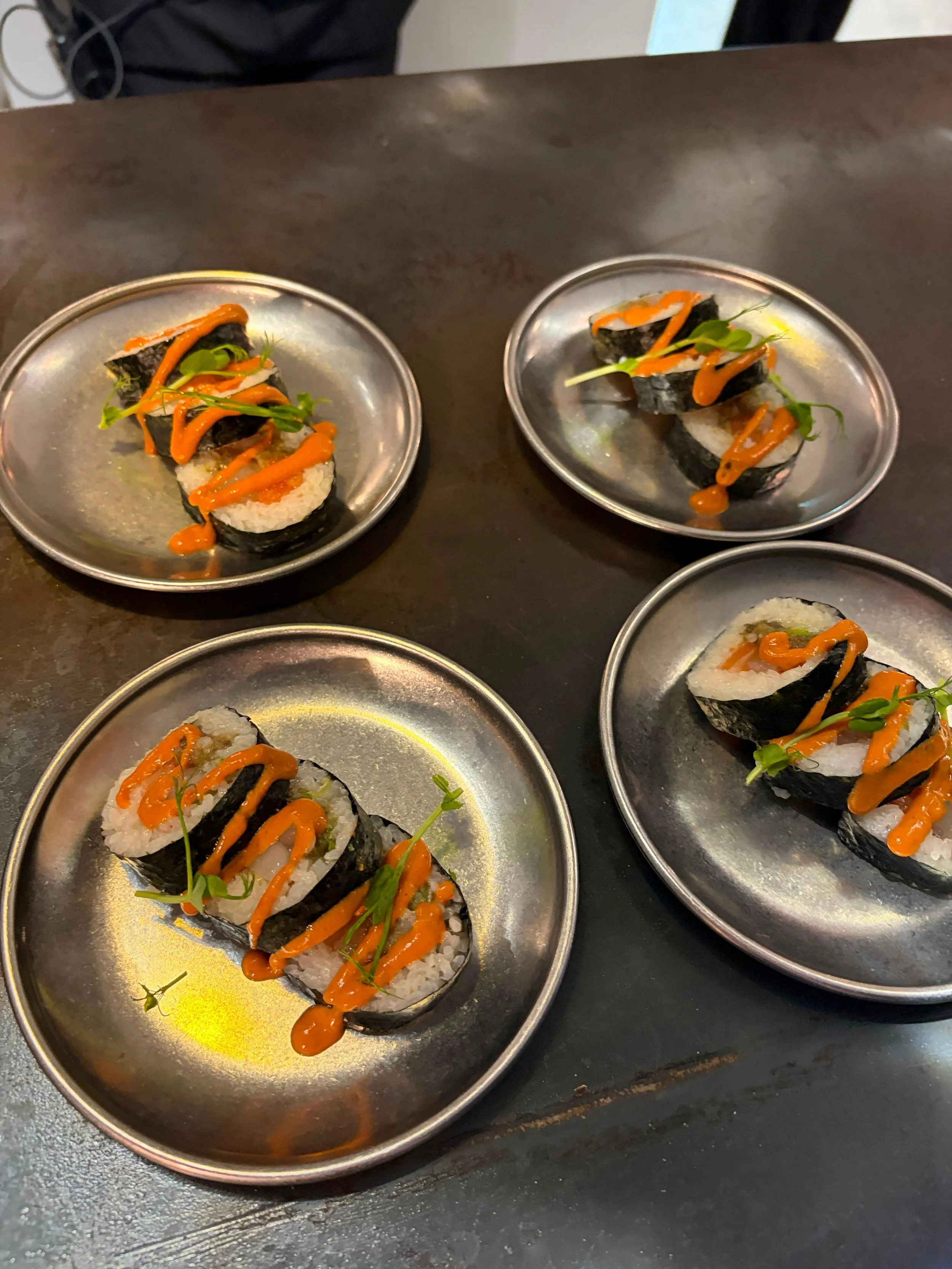Sushi enrollado con arroz, alga nori, salsa y microverdes en platos metálicos.