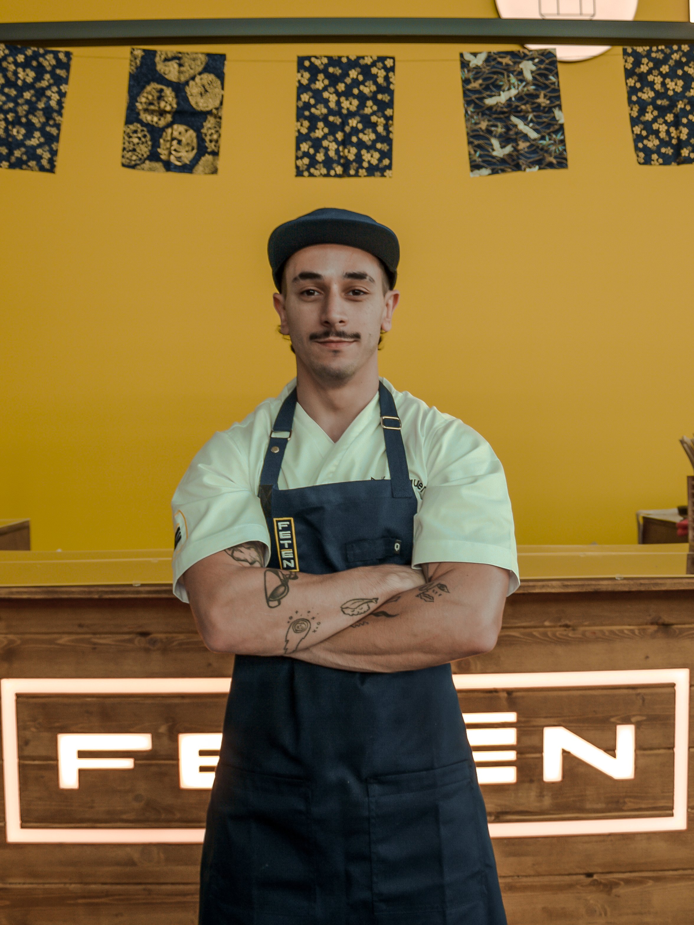 Joven cocinero con barba y tatuajes en brazos, usando uniforme y gorra, de pie frente a un fondo con letras y decoraciones japonesas sobre pared amarilla