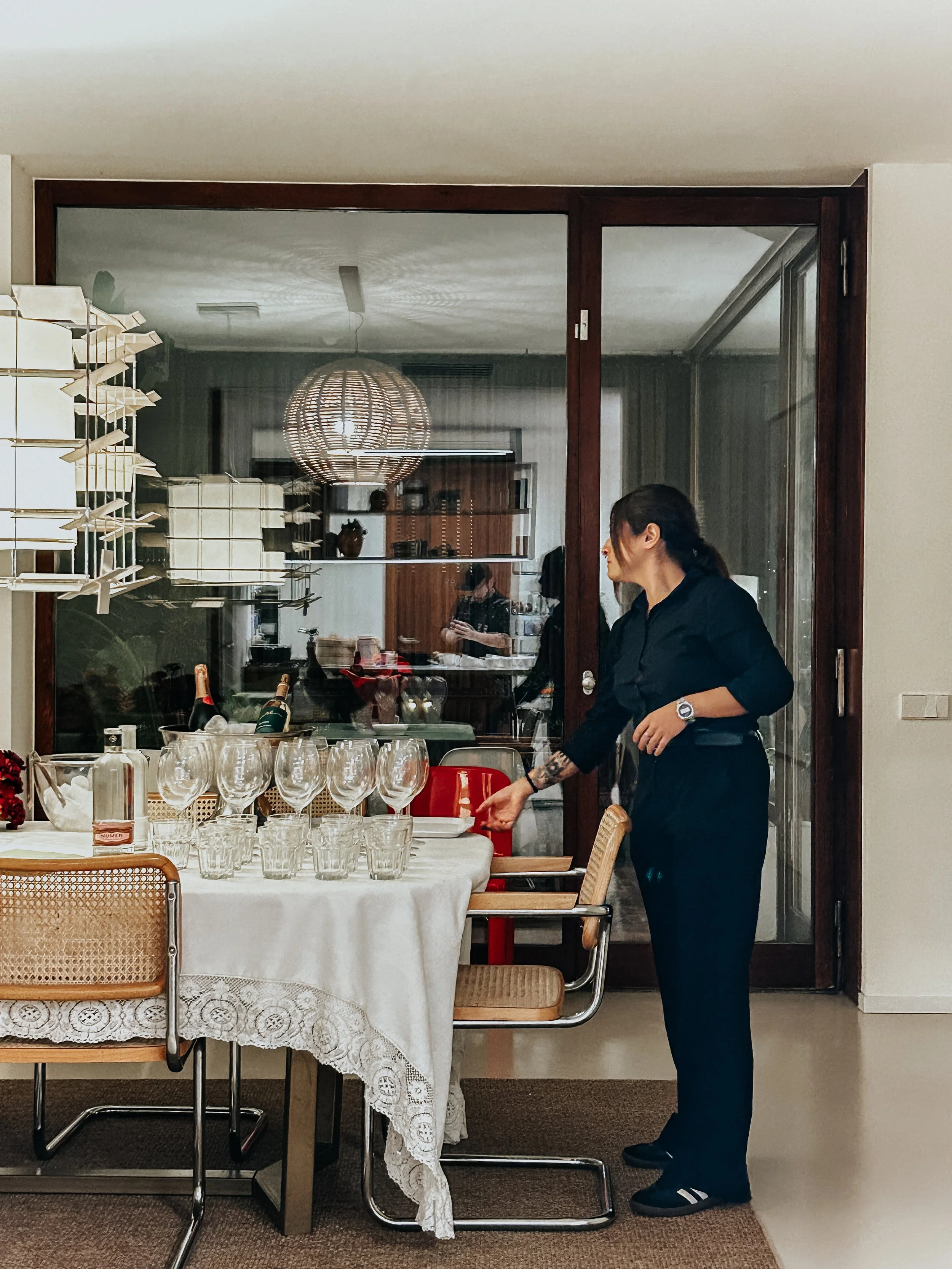 Mujer arreglando copas en una mesa en un comedor, vista a través de un ventanal con reflejo de la luz y decoraciones en el fondo. Servicio de chef privado con camarera. Servicio de bebidas.