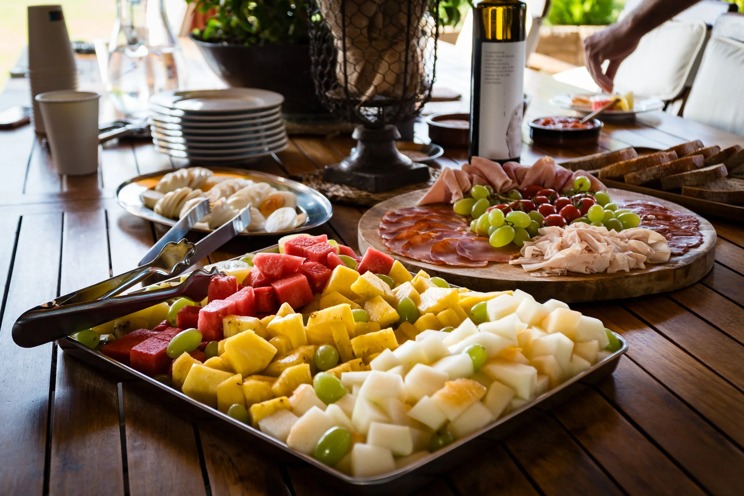 Una mesa de madera con variedad de frutas picadas, jamón, tomates, y otros alimentos, con platos, vasos y utensilios.