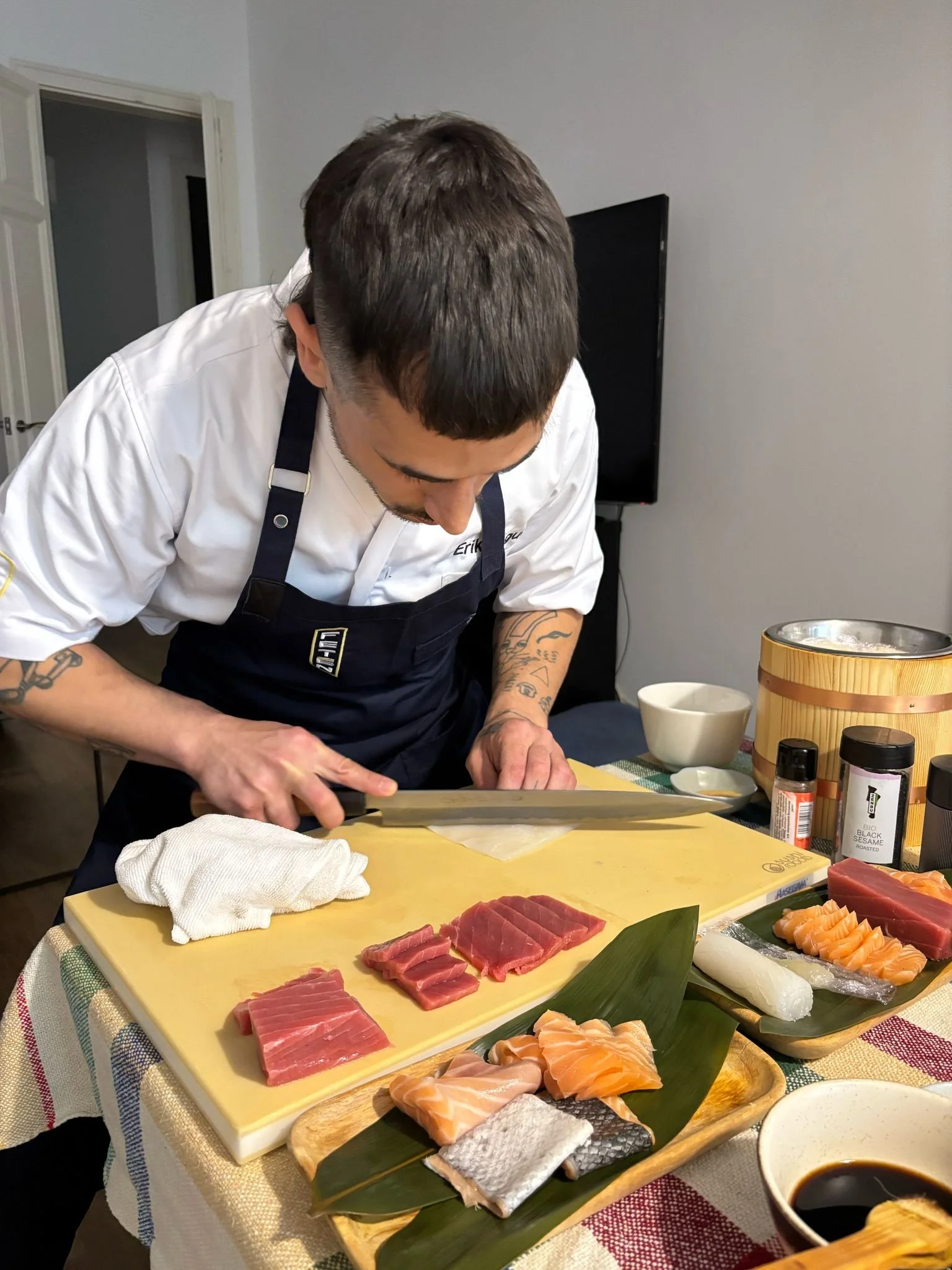 Hombre cortando pescado en una mesa con varios tipos de sashimi y sushi, acompañado de salsa de soja y especias, en un ambiente de cocina o comedor.
