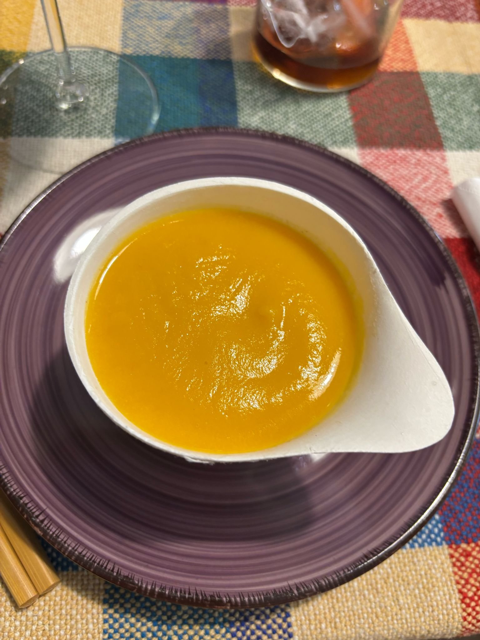 Sopa de calabaza en un plato blanco con borde morado, acompañado de un vaso con bebida oscura y una copa de vino en un mantel de cuadros de colores.