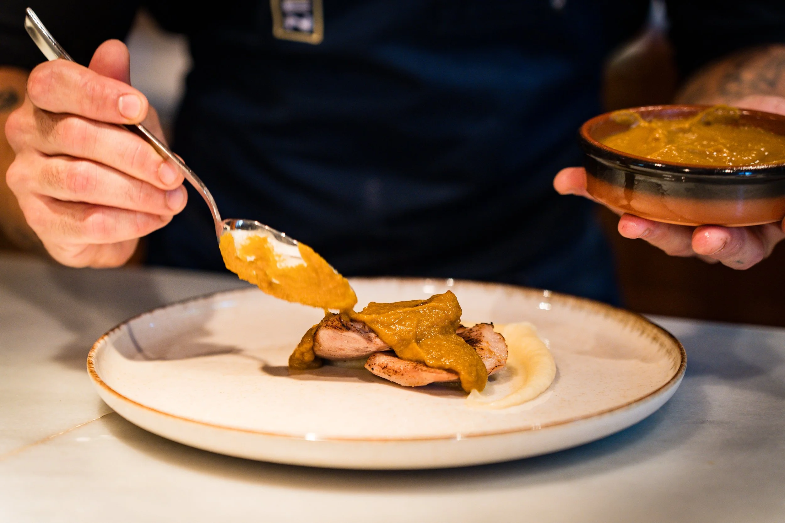 Persona sirviendo carne con salsa sobre un plato con puré y acompañamiento.