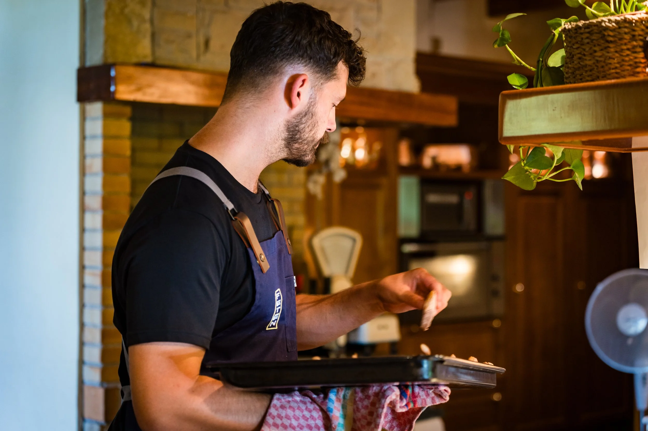 Un hombre con barba y cabello rizado, usando un delantal azul, trabajando en una cocina y sosteniendo una bandeja con alimentos.