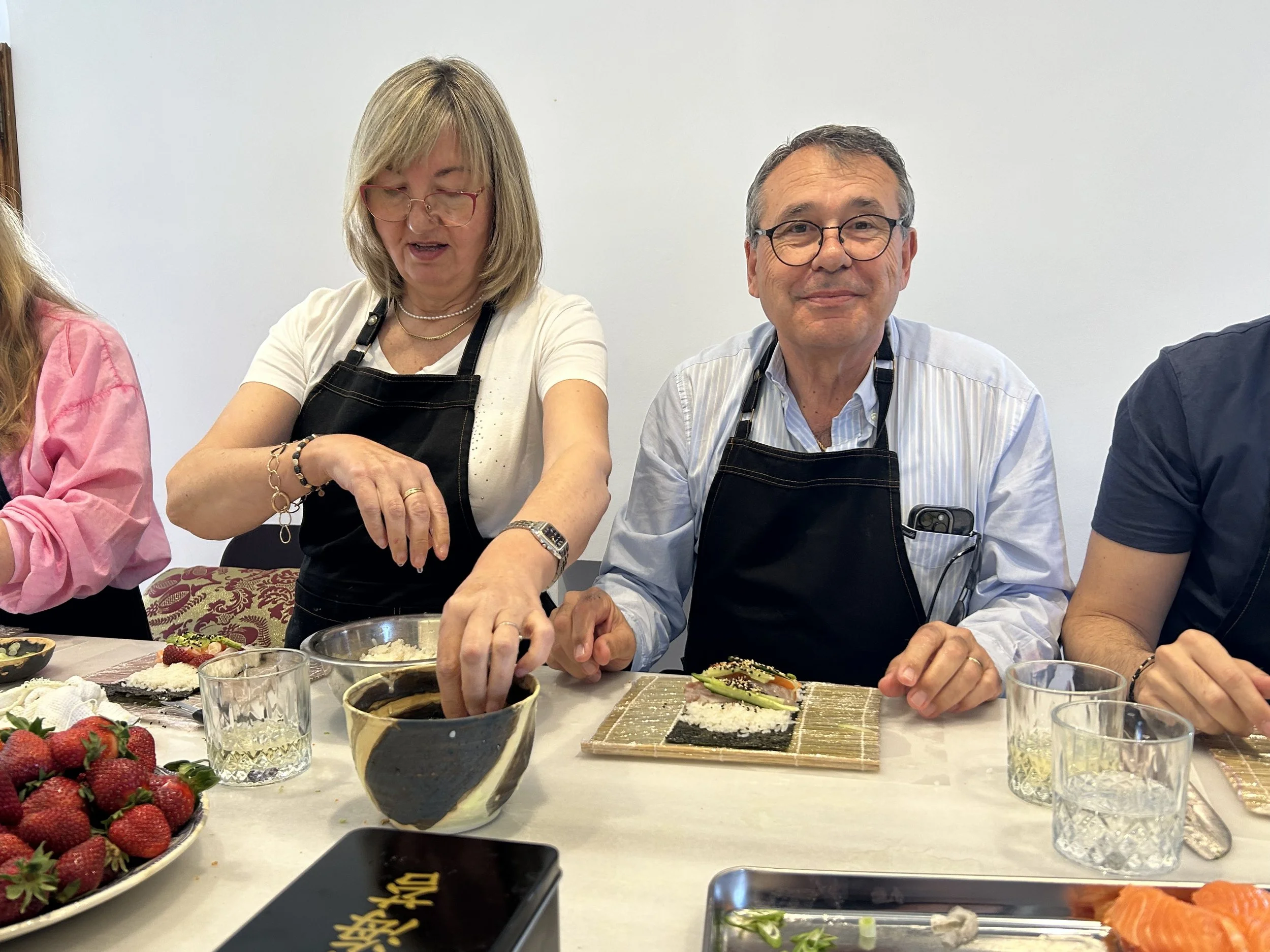 Varias personas sentadas en una mesa disfrutando comida japonesa, como sushi, con frutas, vasos y accesorios en el fondo, probablemente en una reunión o celebración.