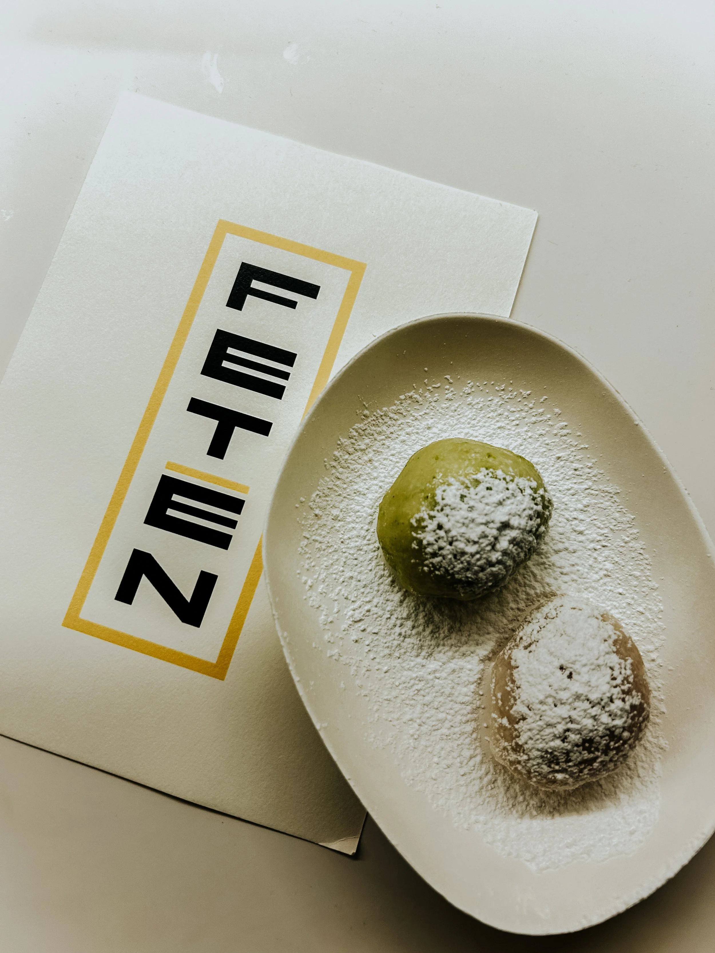 Tres bolitas de masa de color verde, decoradas con polvo blanco, en un plato ovalado. Un sobre con letras en negrita y un borde amarillo es parcialmente visible al lado del plato.