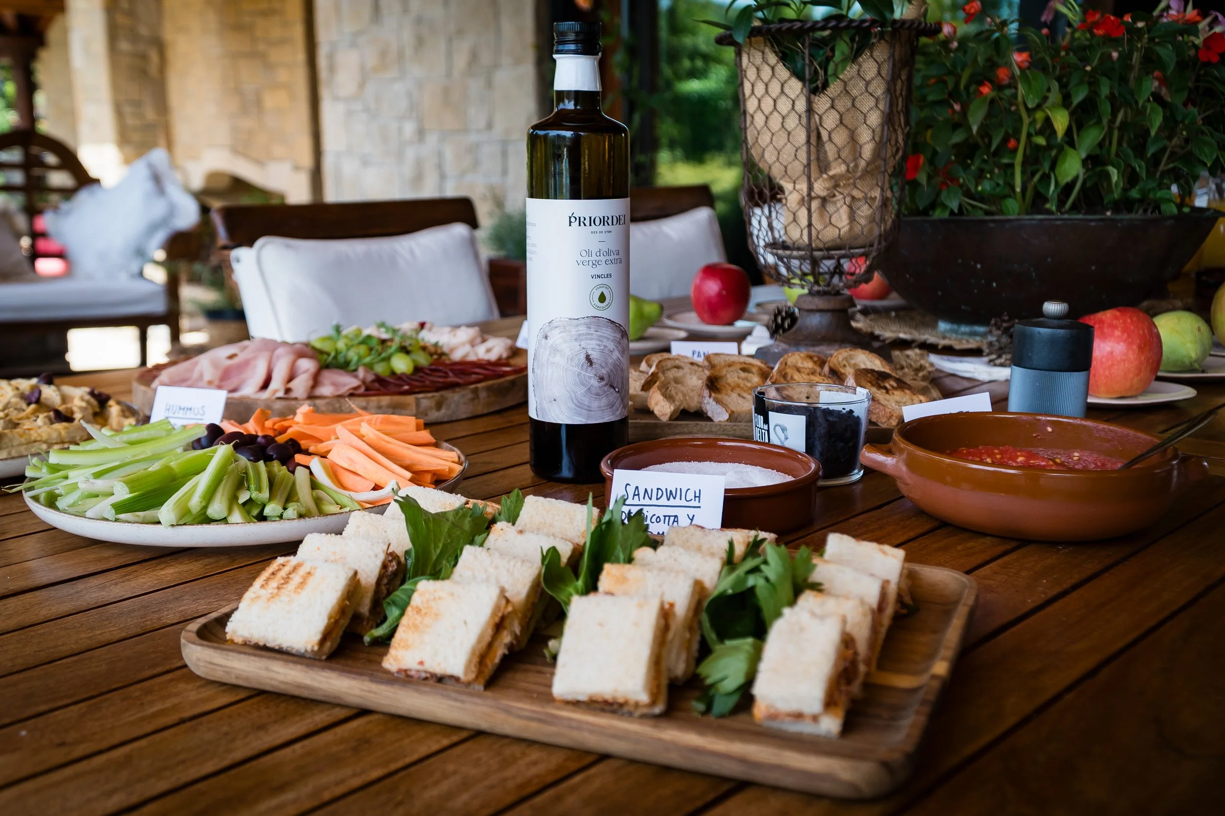 Mesa con alimentos preparados para un picnic al aire libre, incluyendo pan sandwiches, verduras, frutas, y una botella de aceite de oliva. En el fondo hay plantas y un ambiente rústico.