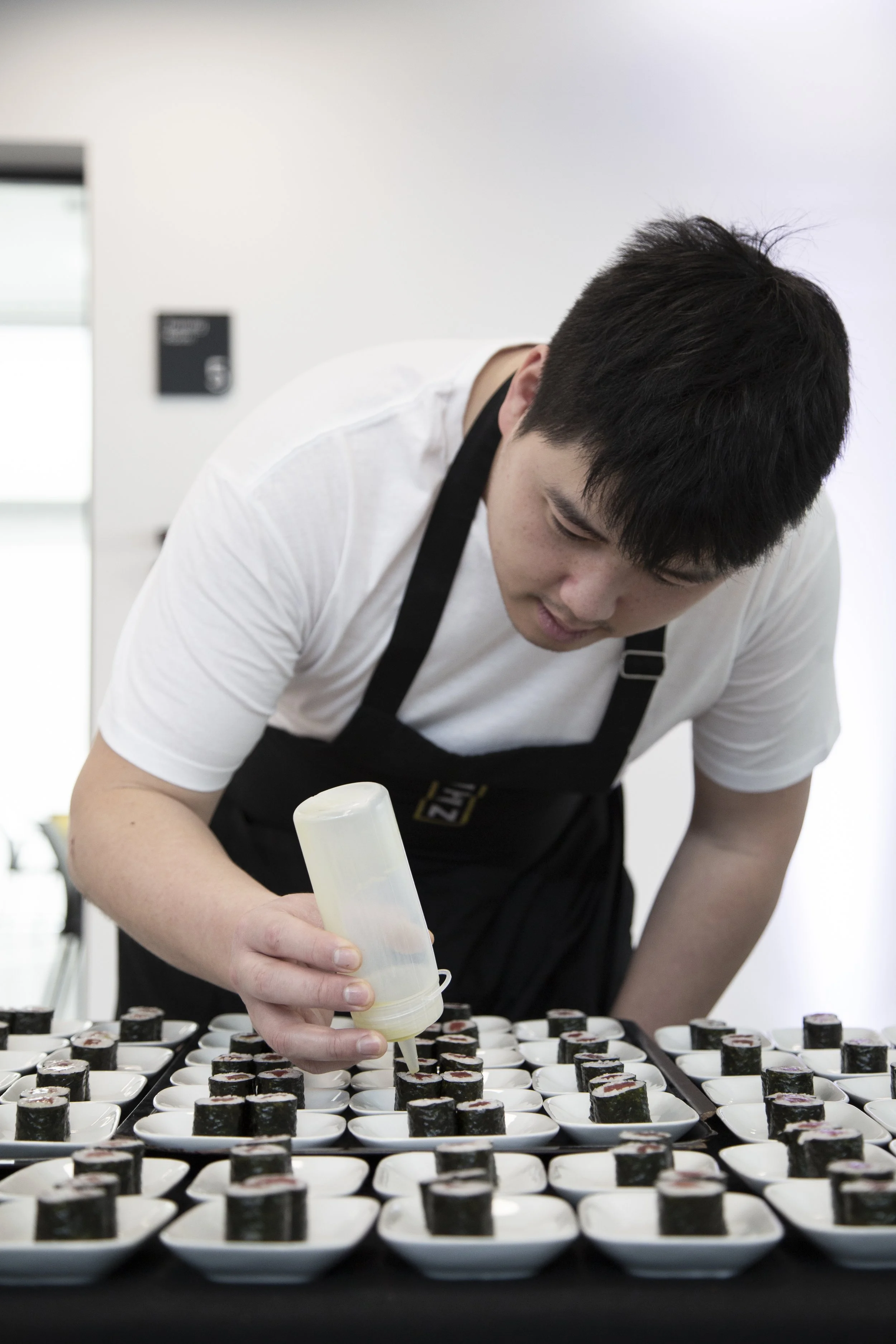 Persona haciendo sushi en una mesa con muchas bandejas pequeñas con sushi y añadiendo salsa. La persona lleva camiseta blanca, delgada y con cabello oscuro. El fondo es blanco con un cartel visible.