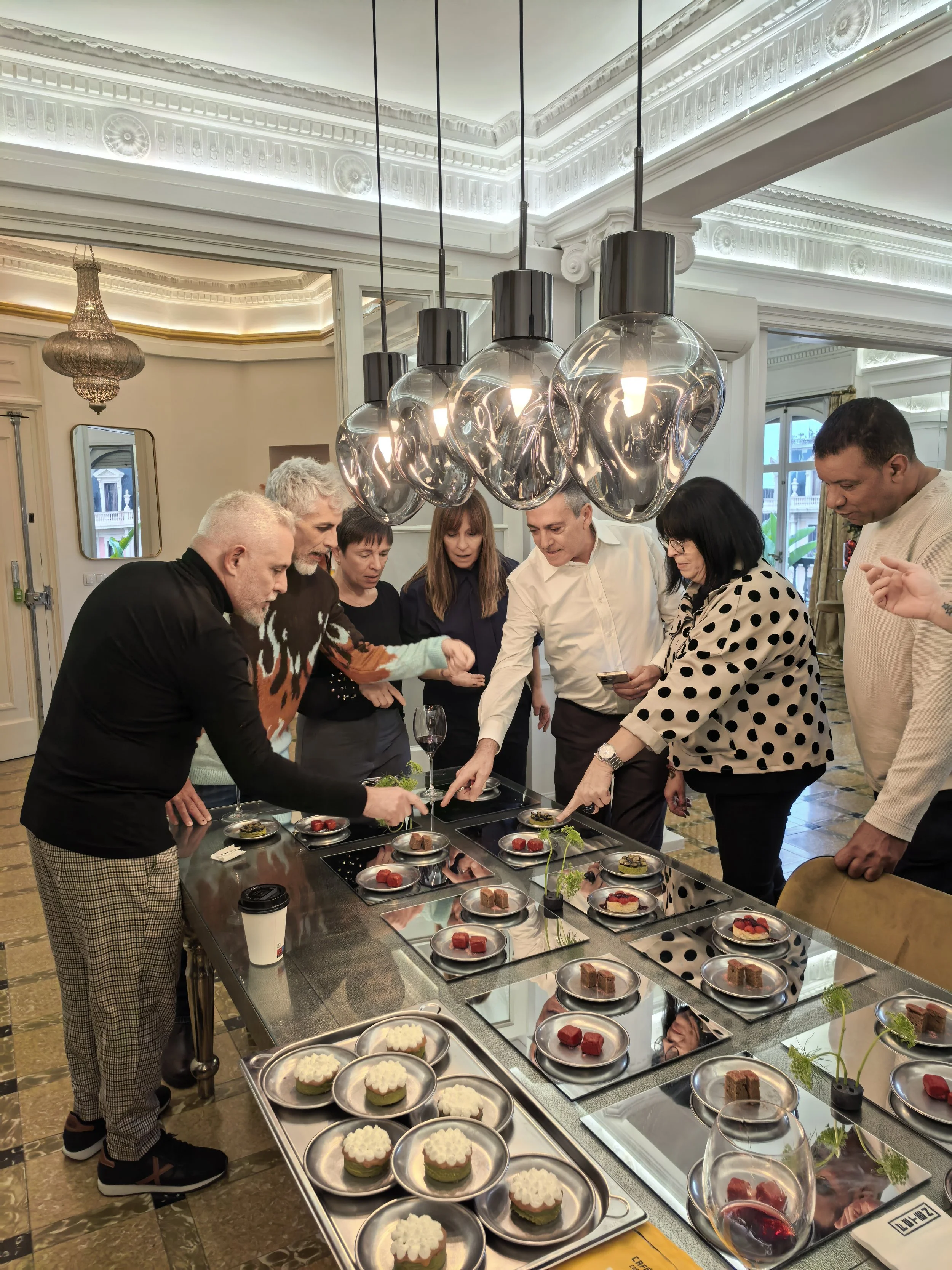 Grupo de personas observando una mesa con postres y cupcakes en un entorno elegante con iluminación moderna y decoraciones clásicas.