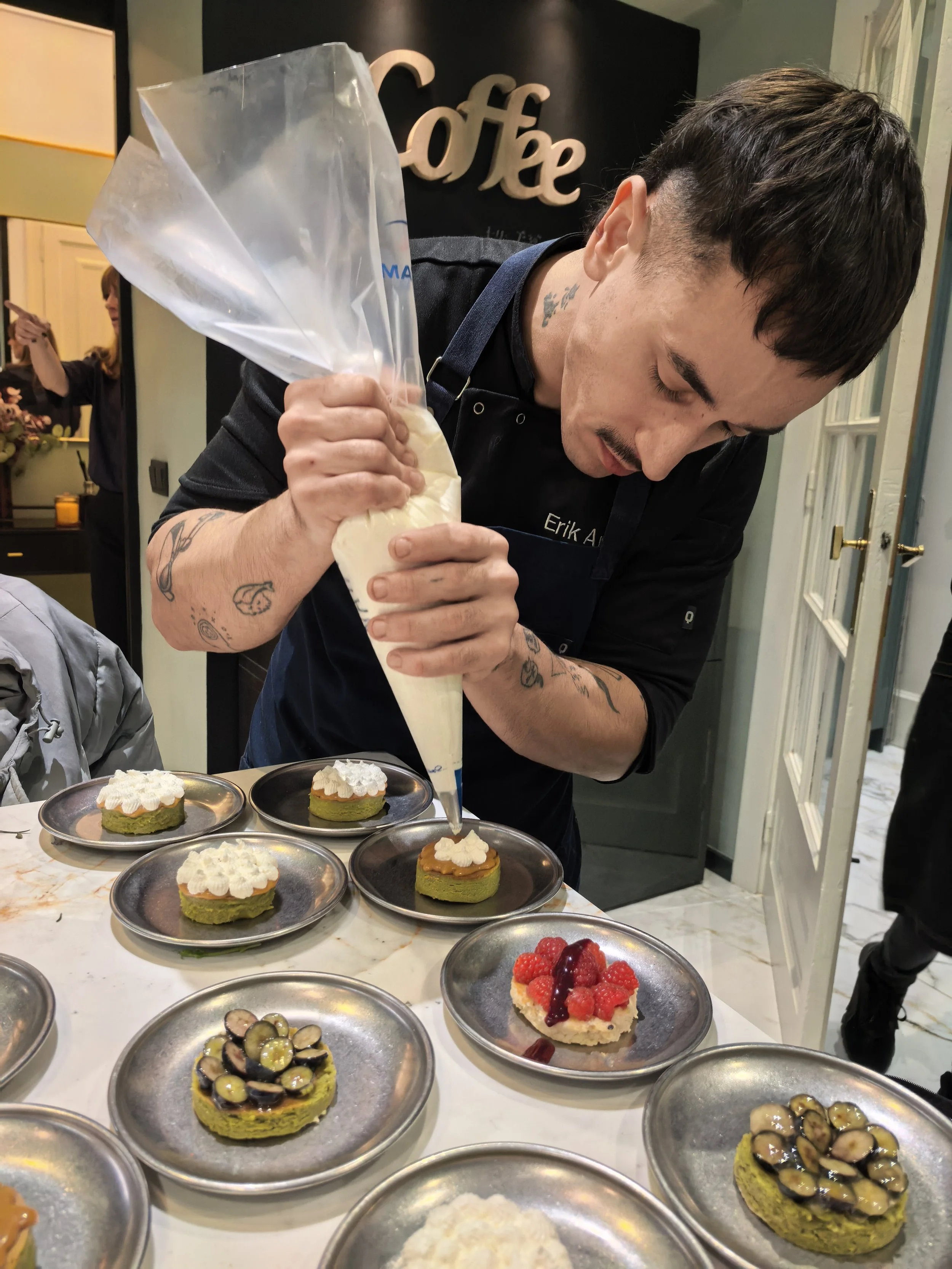 Un hombre decorando postres con crema en una cafetería, rodeado de platos con diferentes postres y frutas.