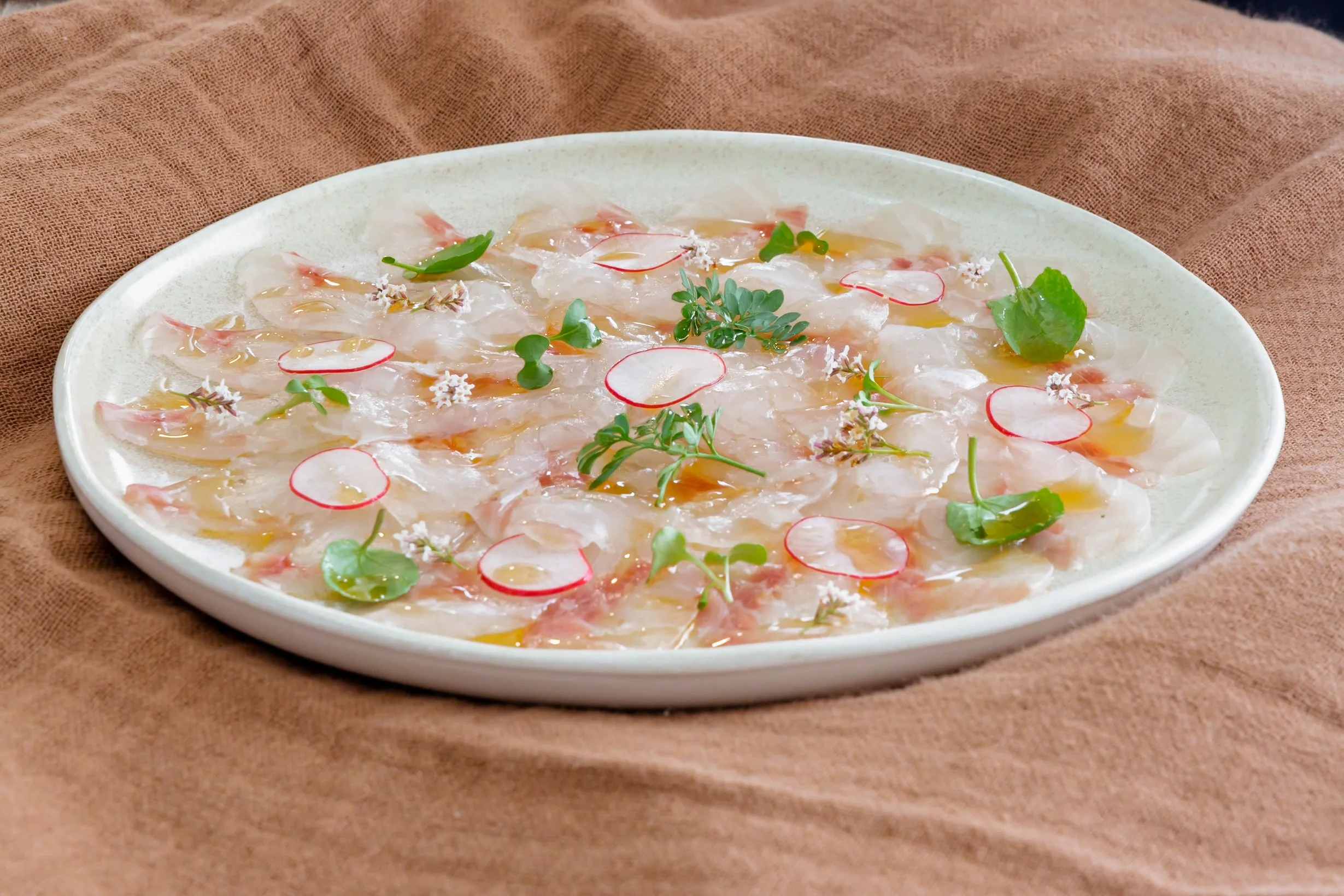 Plato de carpaccio de pescado con finas rodajas, adornado con hierbas frescas, rodajas de rábano y un poco de aceite o salsa