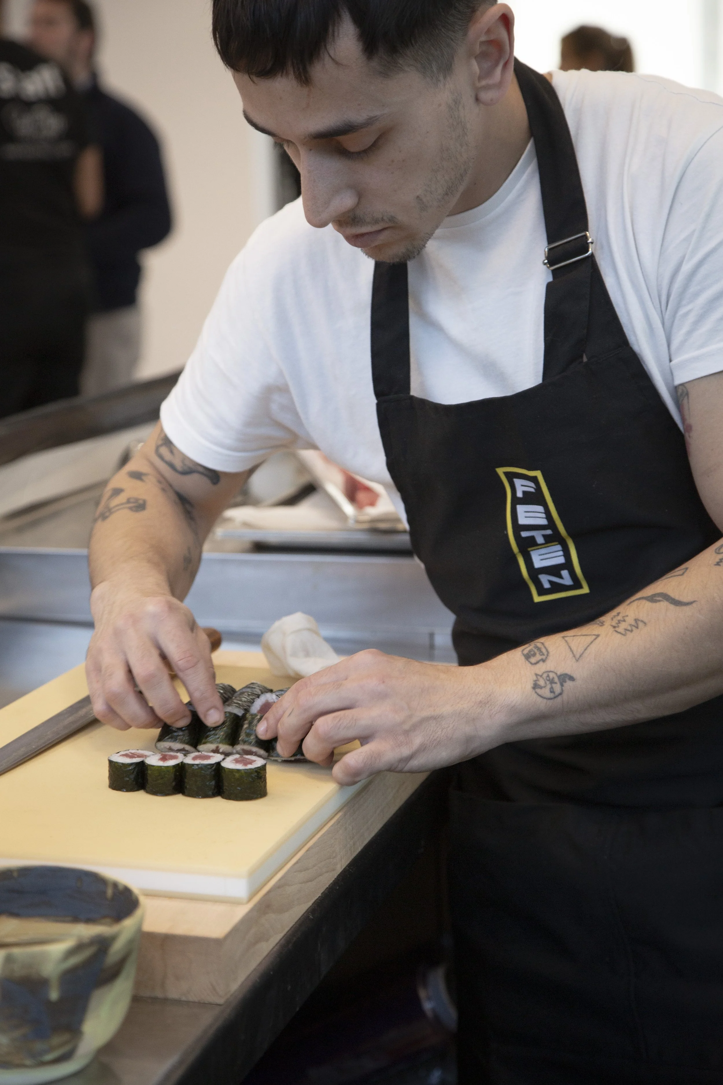 Un joven chef prepara sushi en una mesa, cortando rollos de sushi con una cuchilla. Tiene tatuajes en los brazos y lleva una camiseta blanca y un delantal negro.