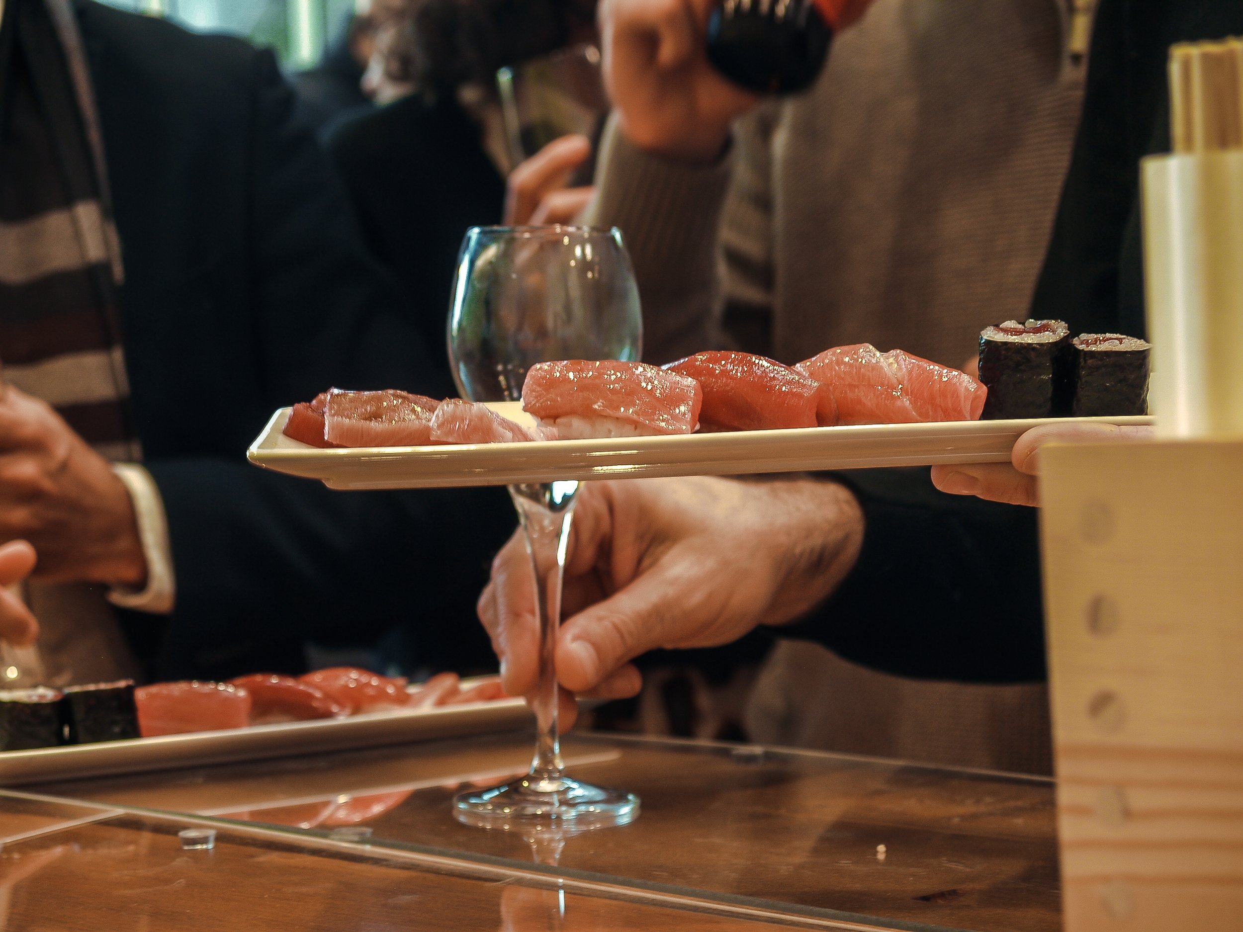 Persona sirviendo sashimi de atún en una copa de cóctel en un restaurante japonés, con otros comensales al fondo.