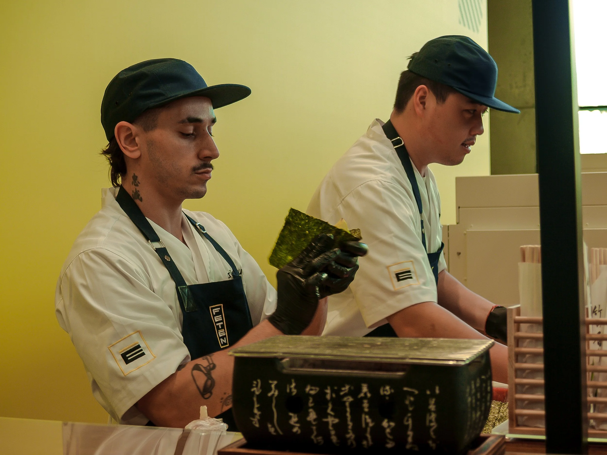 Dos hombres con delantales blancos y gorras negras trabajan en una cocina o mostrador, uno de ellos sostiene una hoja de alga nori.
