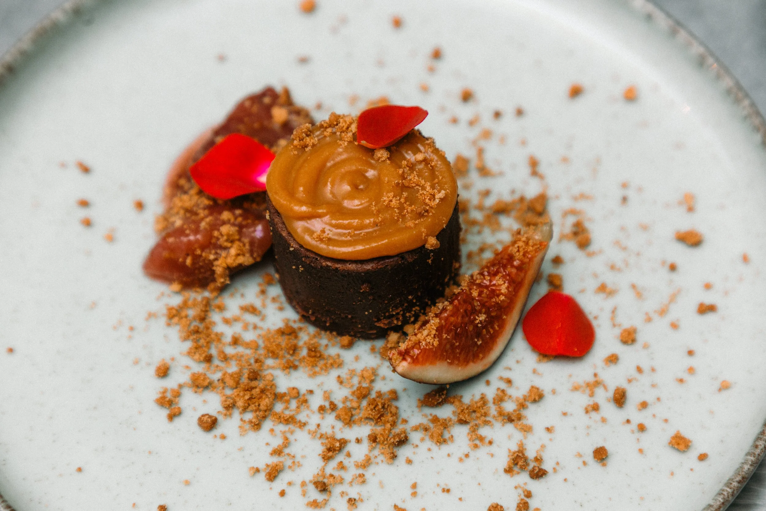 Postre con base de chocolate, decorado con caramelo, pétalos de rosa y polvo de galleta en un plato blanco.