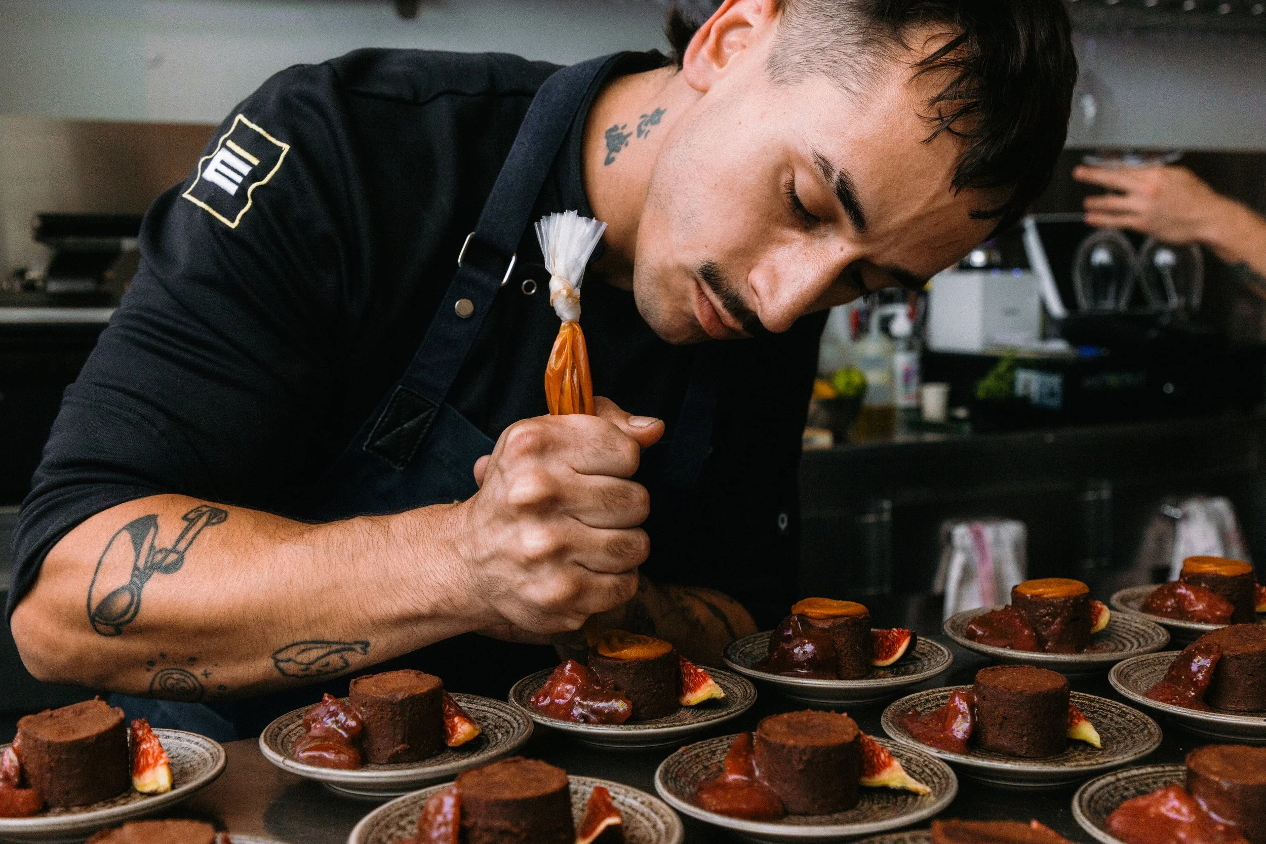 Hombre decorando postres en una cocina, rodeado de platos con postres de chocolate y frutas.