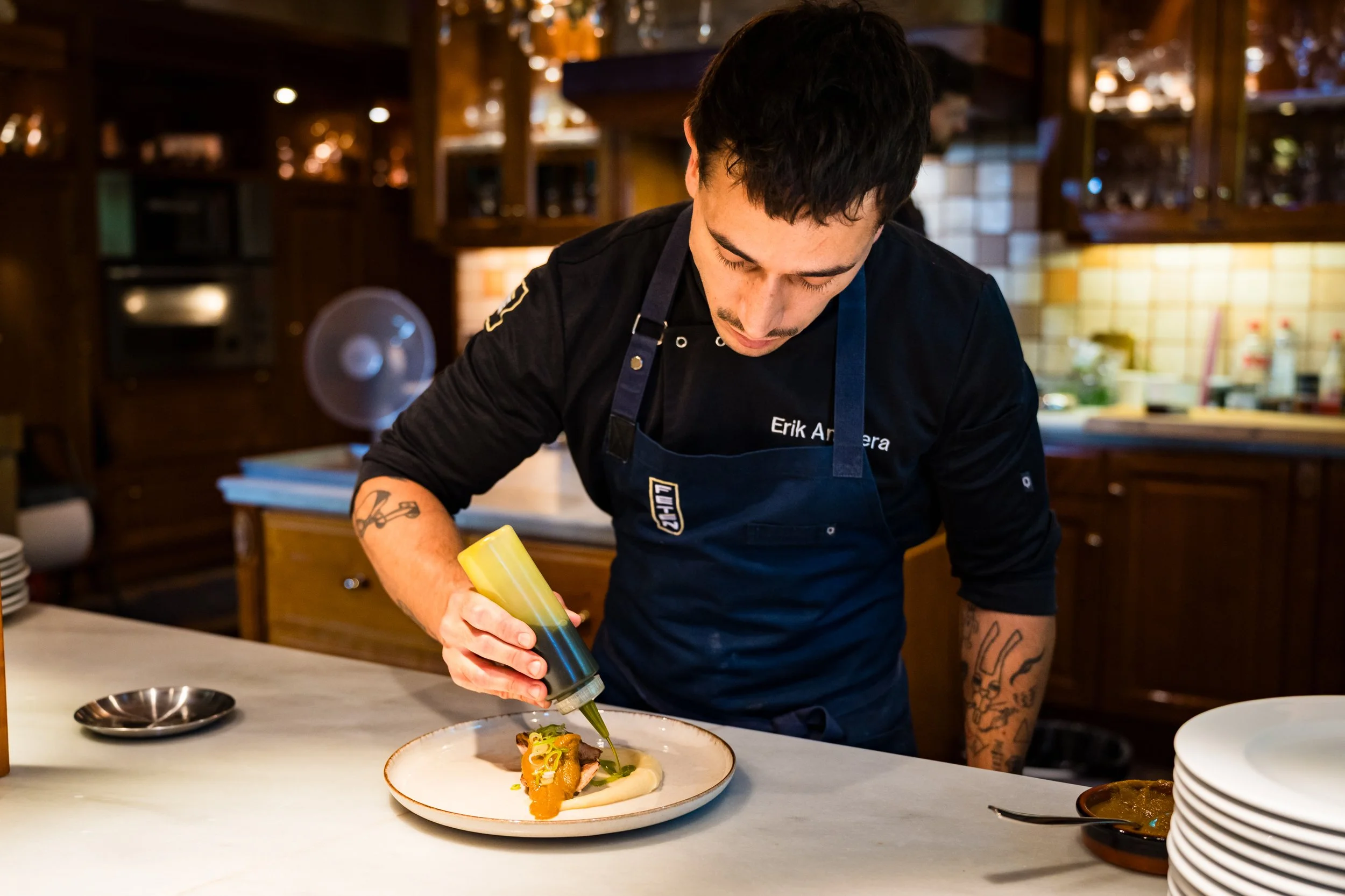 Un chef decorando un plato con salsa desde una botella en una cocina rústica, con utensilios y platos apilados en el fondo.
