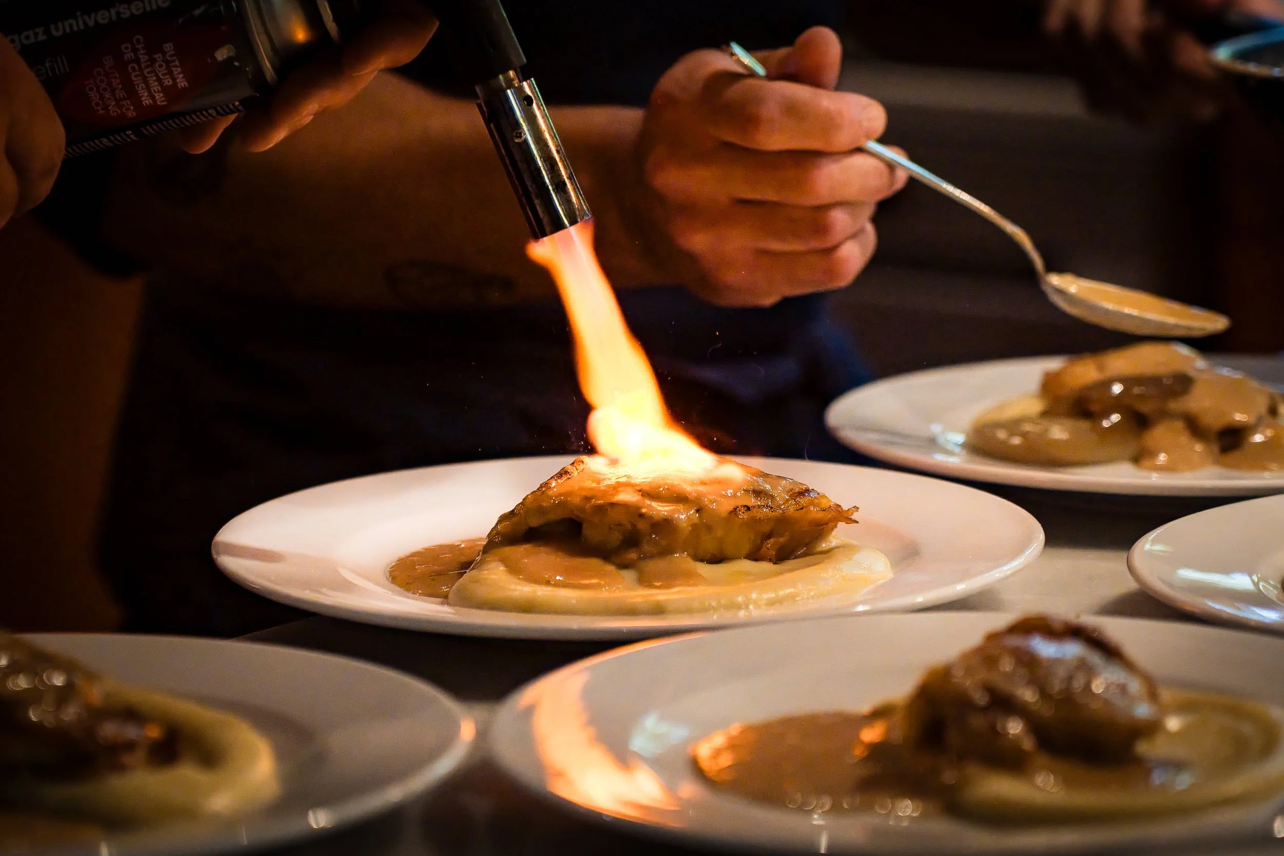 Persona usando un soplete de cocina para caramelizar un postre en un plato. Hay otros platos similares en la mesa.