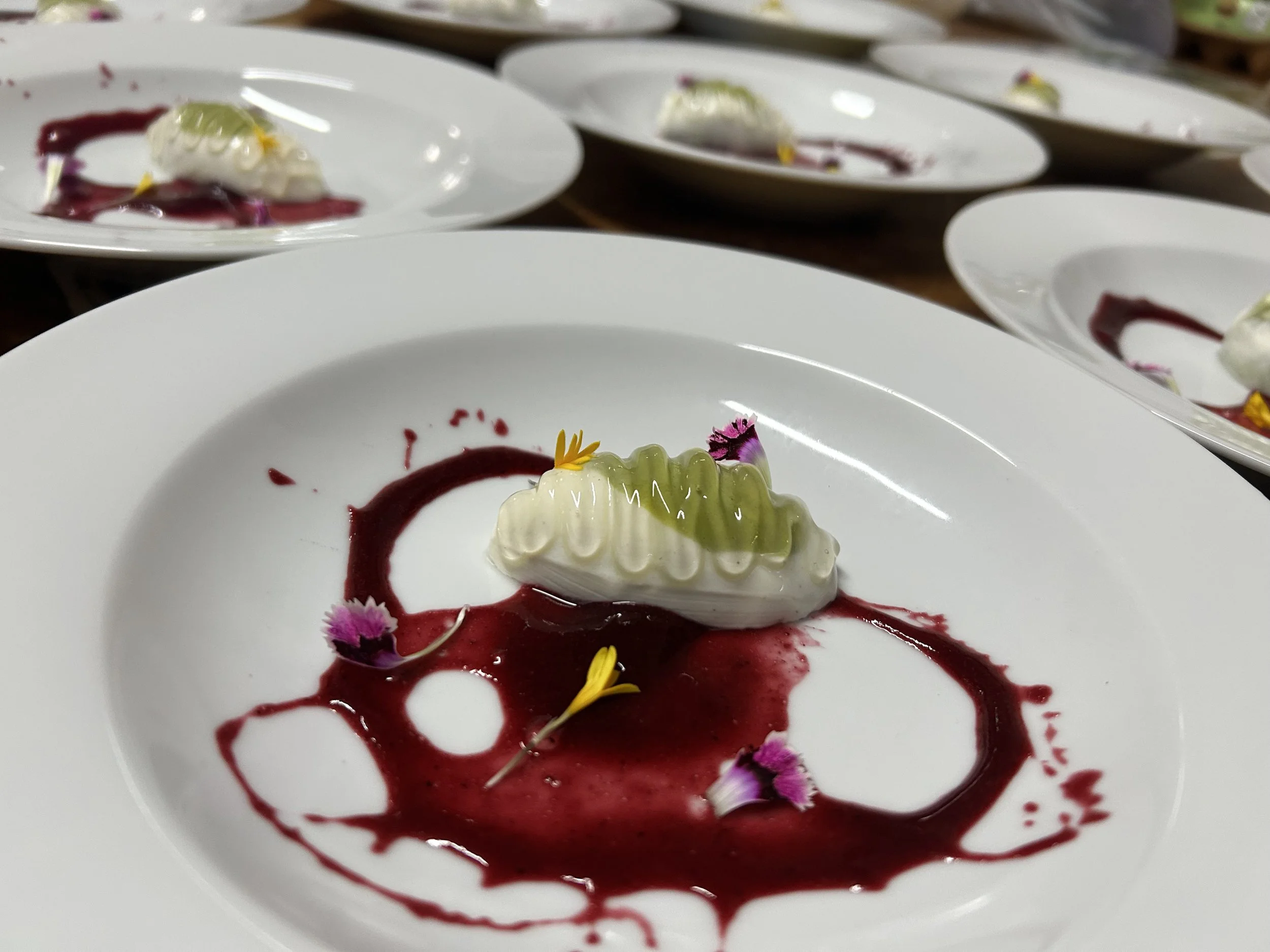 Postre con crema, salsa de frutos rojos, flores y decoraciones comestibles en platos blancos.