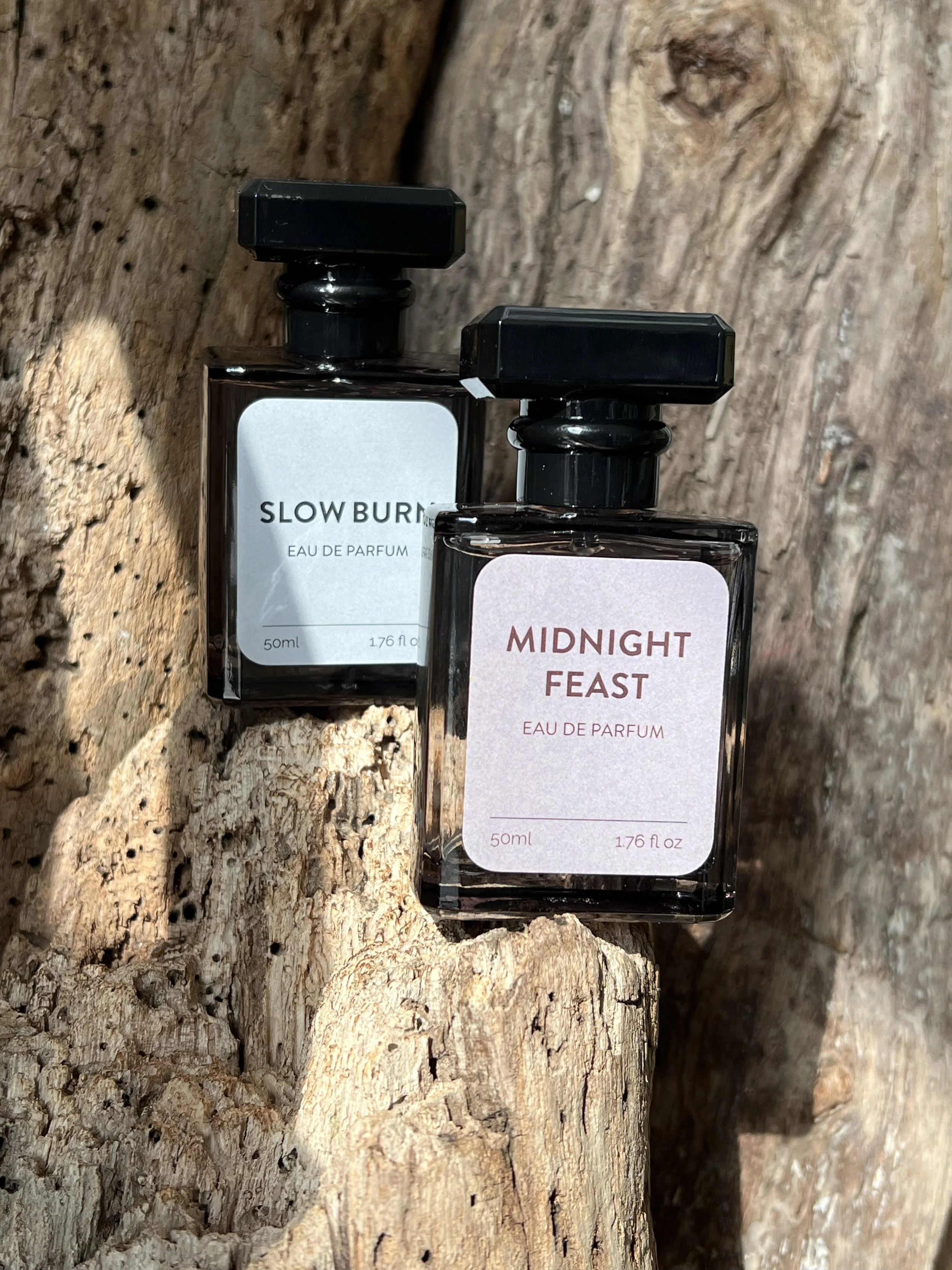 Two perfume bottles placed on a rough wooden surface. The bottle on the left is labeled "SLOW BURN" and the one on the right is labeled "MIDNIGHT FEAST." Both bottles are rectangular with black caps.