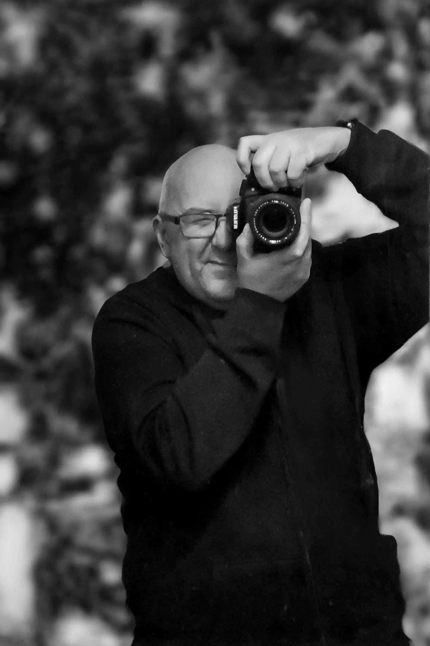 A man taking a mirror selfie with a camera, wearing glasses and a black long-sleeve shirt, with a blurred outdoor background.